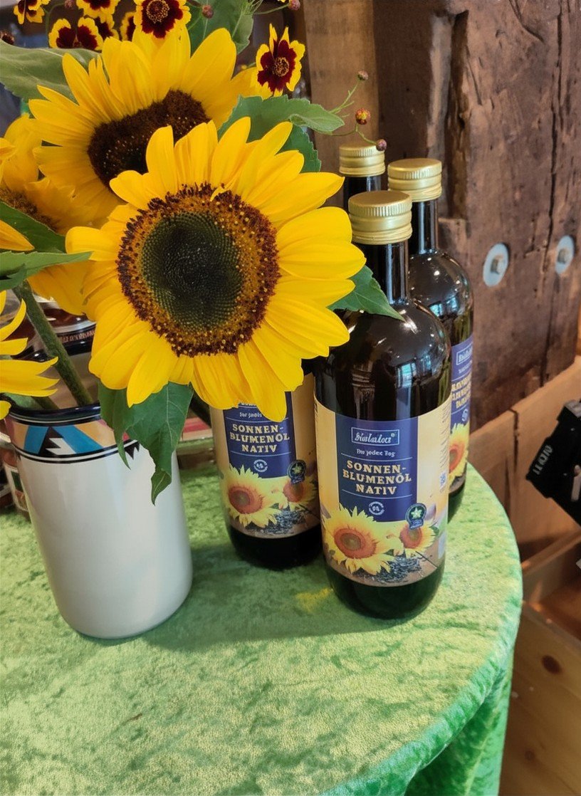 Vasen mit Sonnenblumen und anderen Blumen neben Flaschen mit Sonnenblumenöl auf grünem Tuch vor Holzwand.