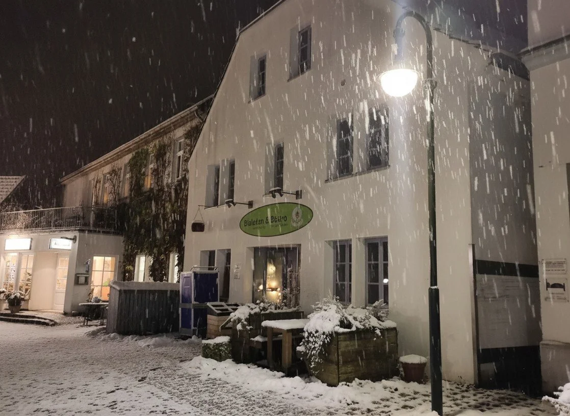 Schneegestürmte Straße vor einem weißen Gebäude mit Beleuchtung, Schneefall sichtbar.