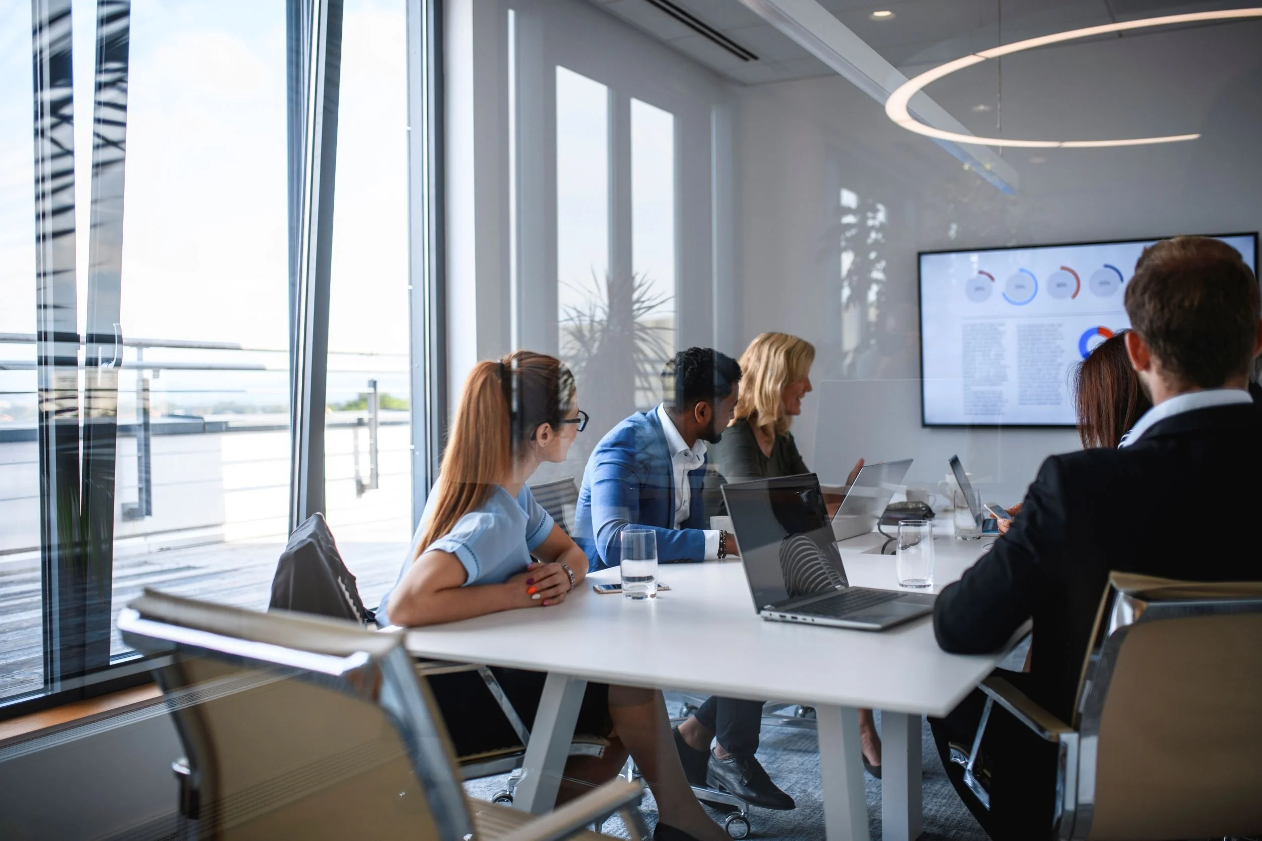 Business professionals attending a meeting in a modern conference room with large windows, laptops on the table, and a screen displaying graphs and data.
