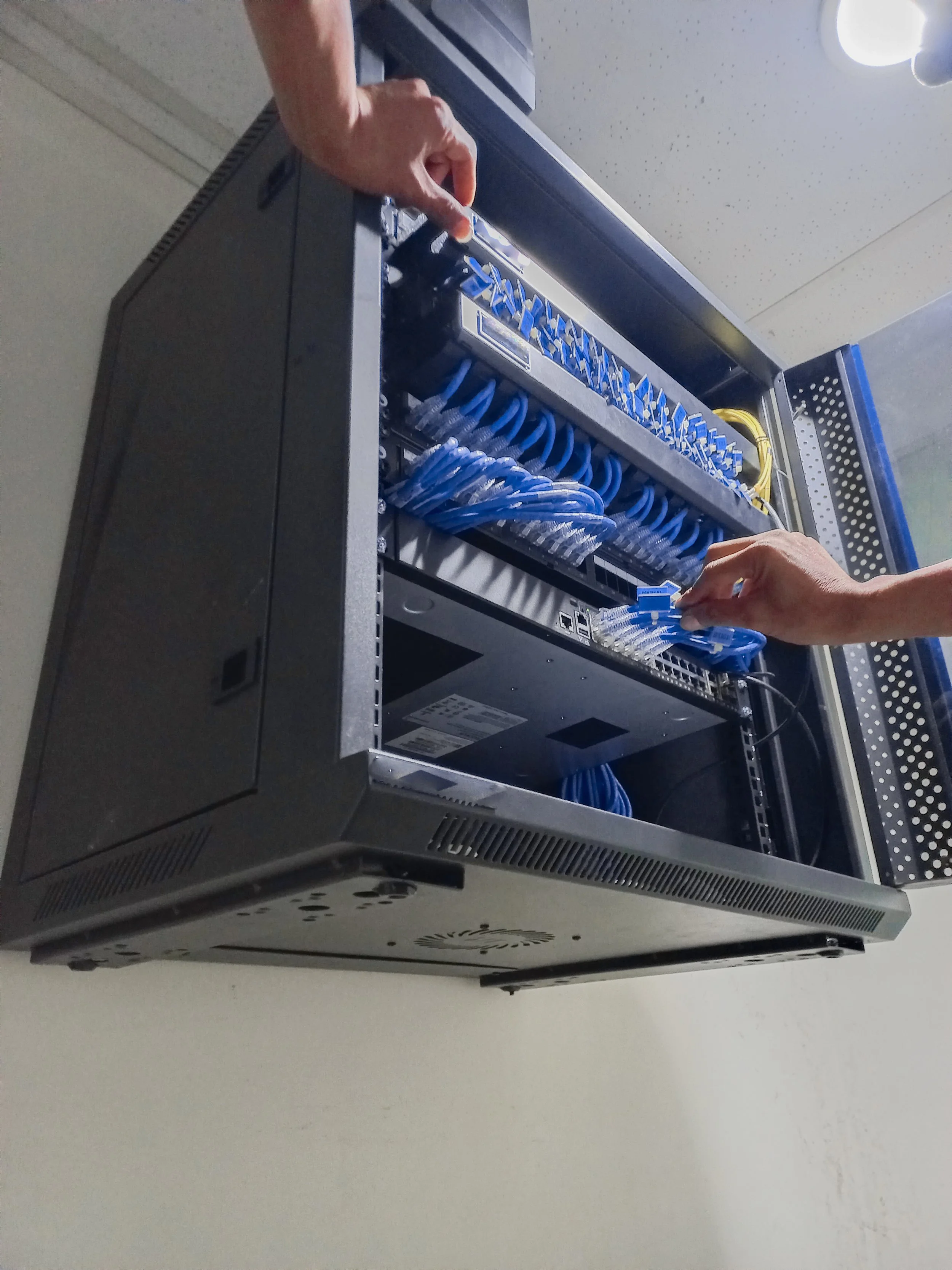 Two hands working on assembling or maintaining a network switch or server with multiple Ethernet cables connected to it, inside a metal rack.