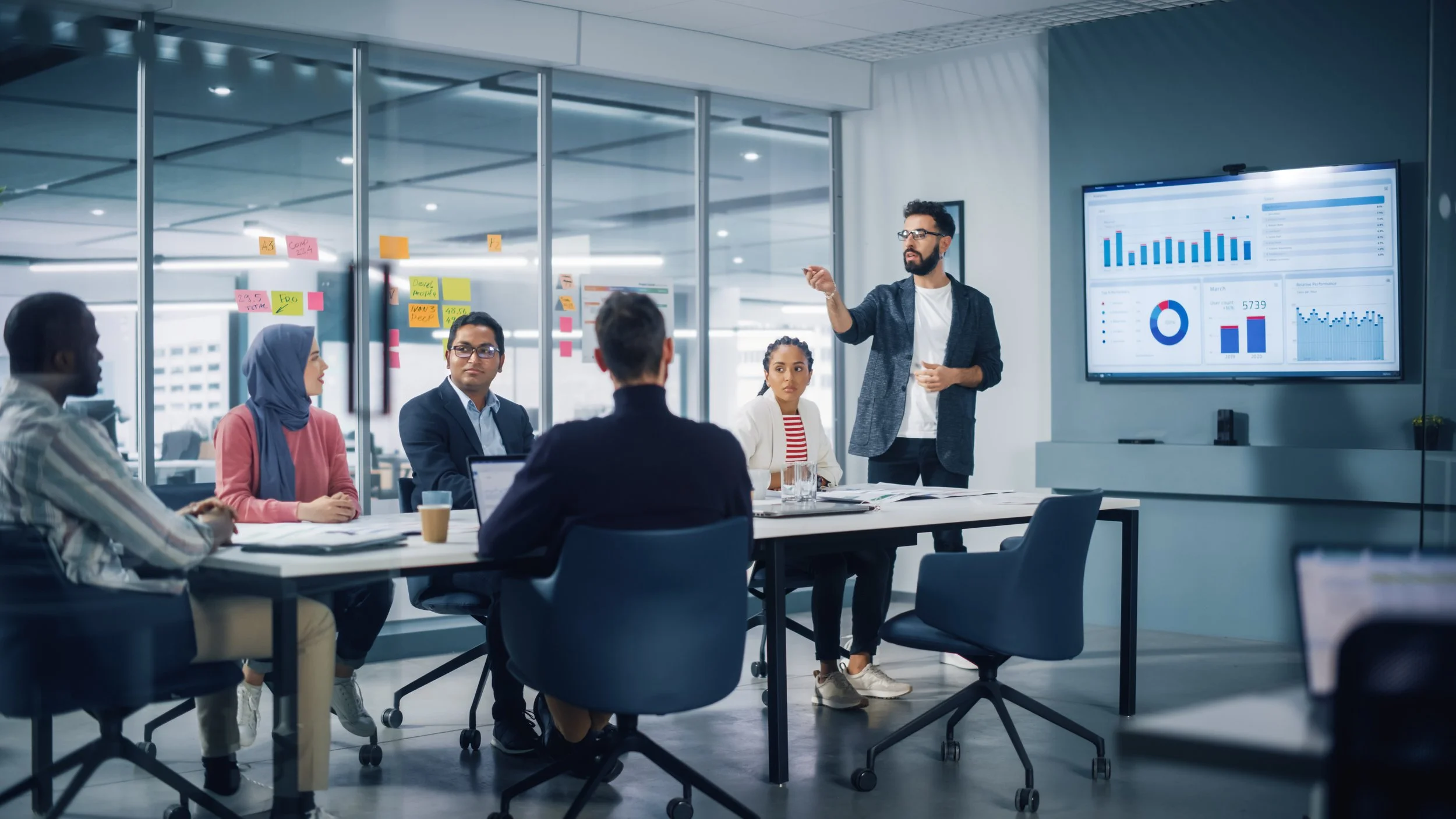 A man presenting data to a group of diverse professionals in a modern conference room with a large digital screen displaying charts and graphs.