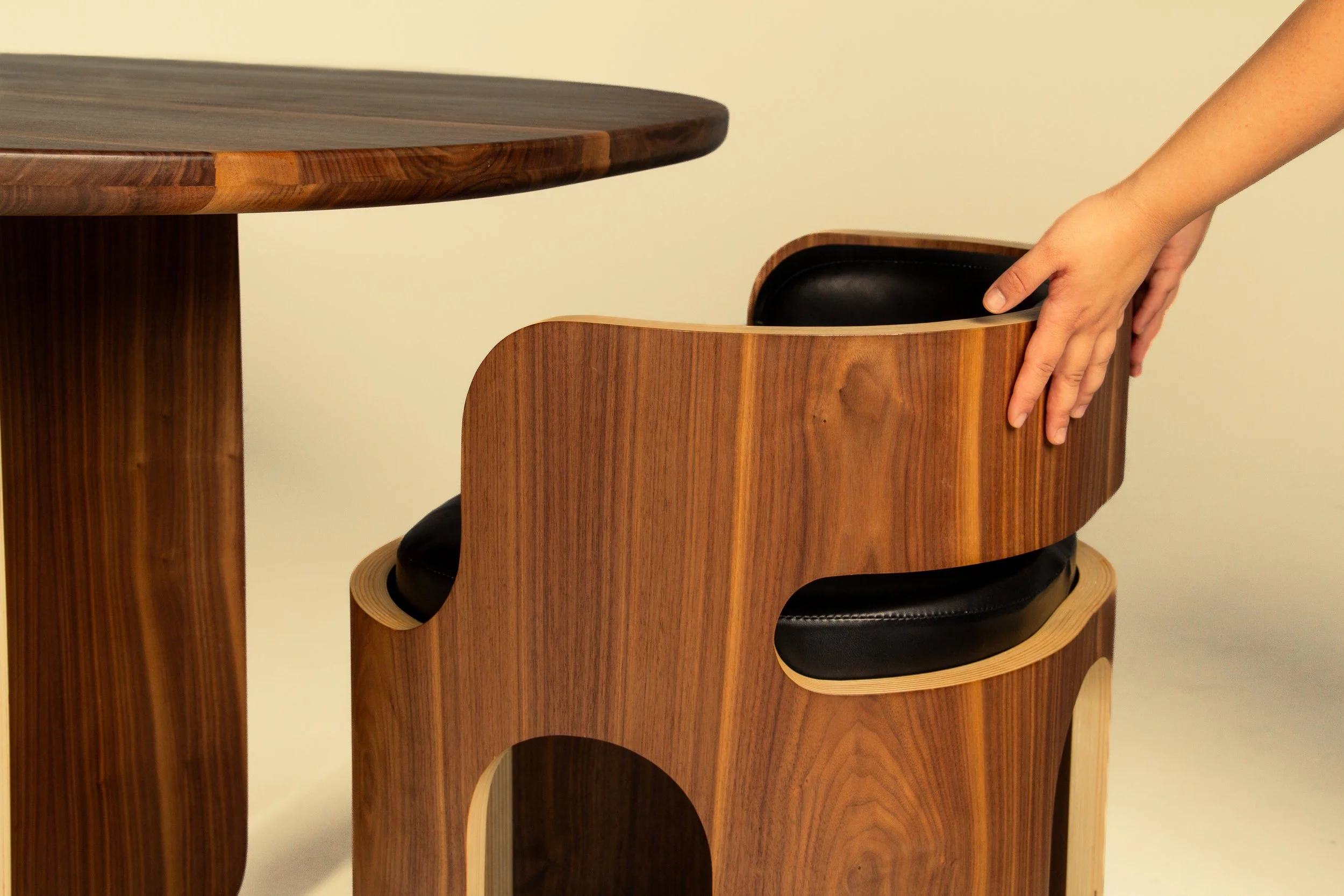 A person is placing a wooden chair with black cushions underneath a wooden table, showing modern furniture design.