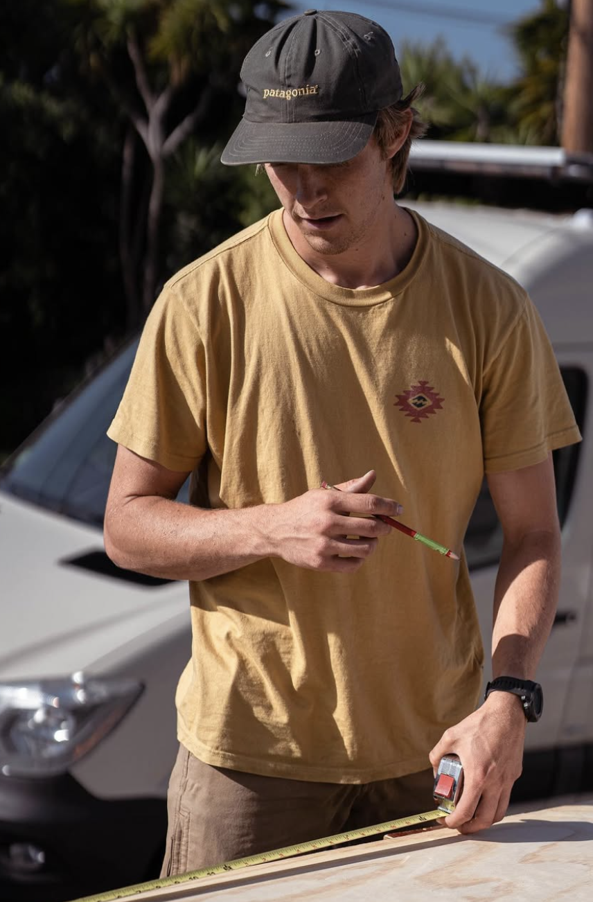 A young man in a yellow t-shirt and gray Patagonia cap measures and marks a piece of wood with a tape measure and a pencil outdoors, with a car and trees in the background.