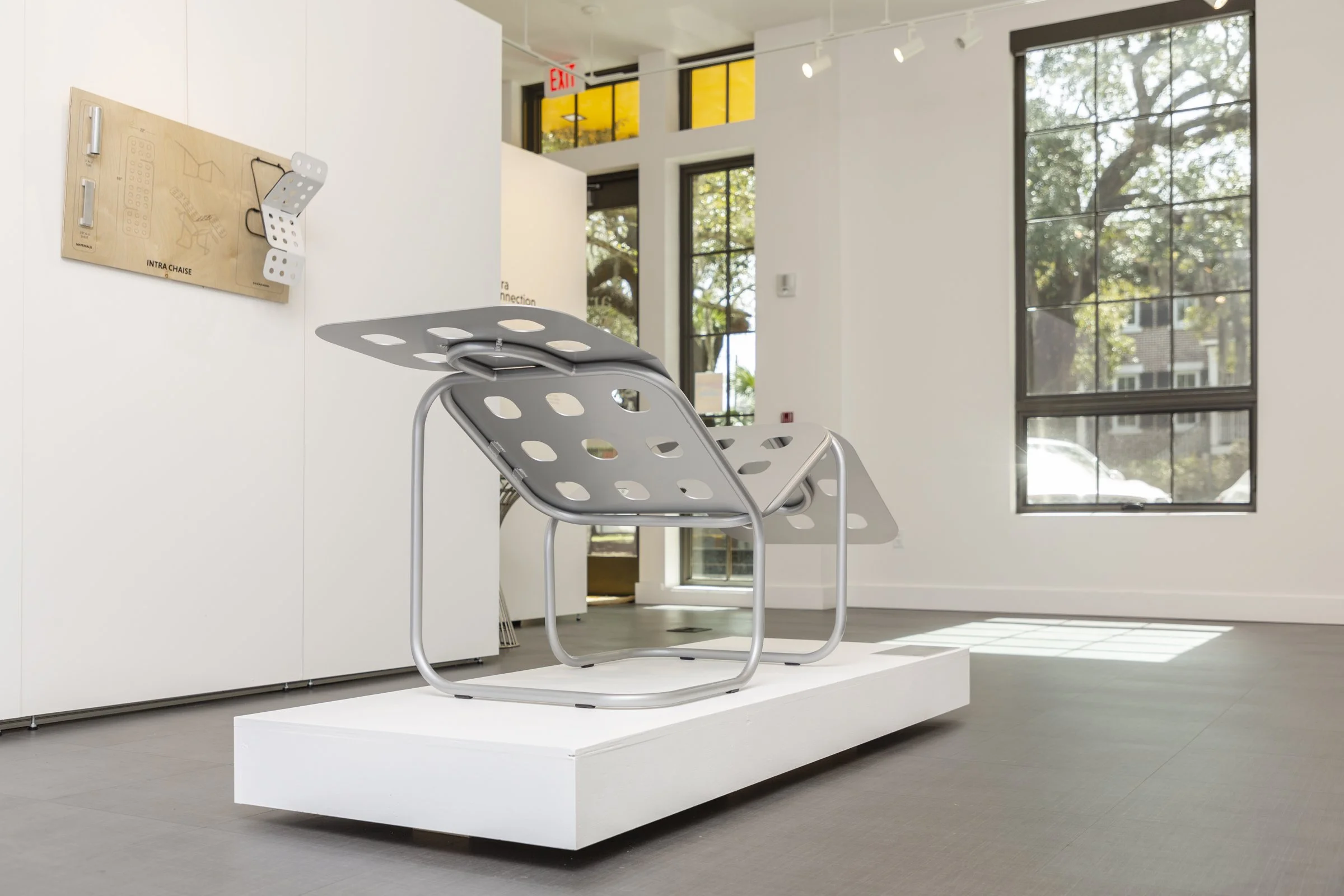 A modern art installation featuring a metal chair with large holes and a flat top, placed on a white platform in a gallery space with large windows and white walls.
