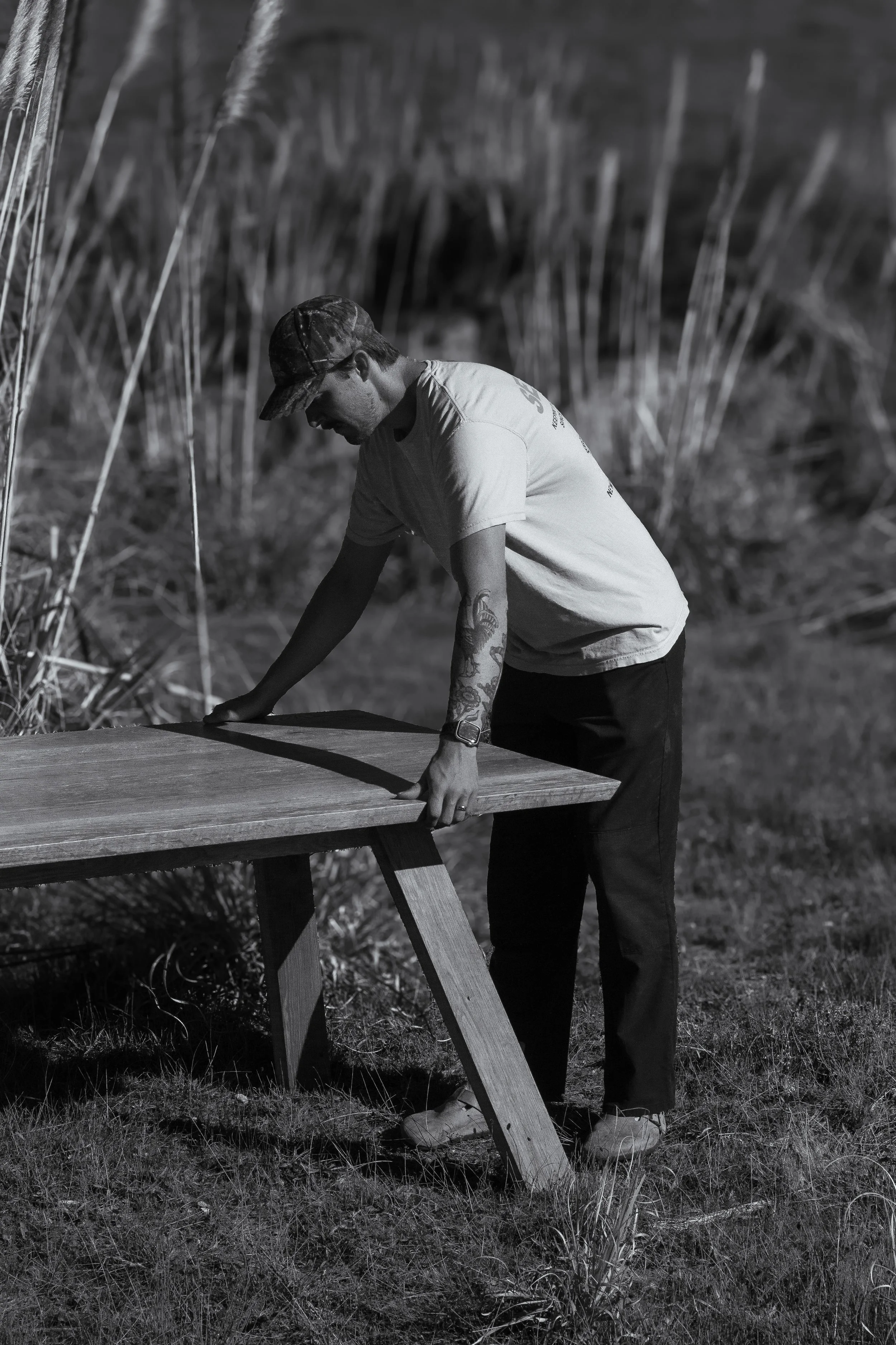 A man with tattoos and a cap assembling a wooden table outdoors in a grassy area with tall reeds in the background.