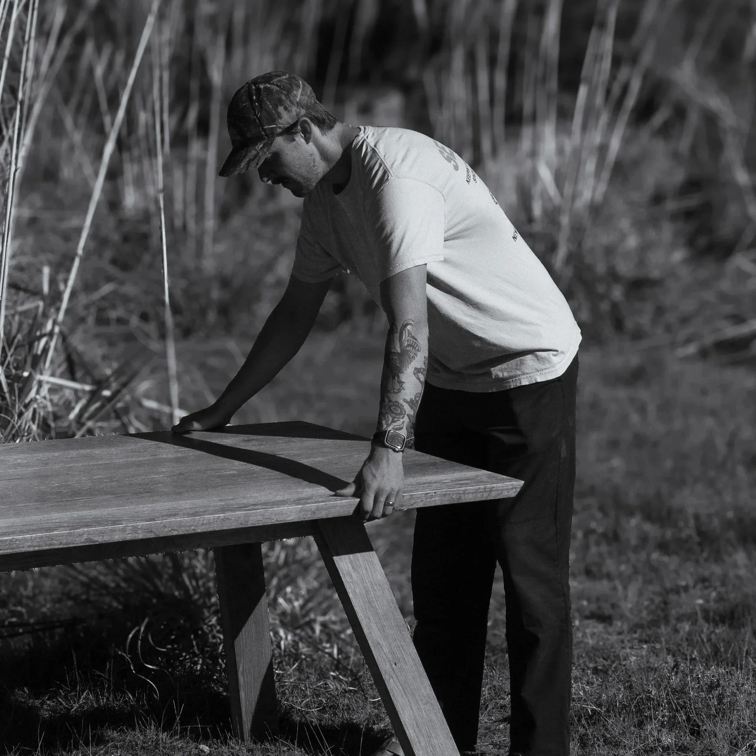A man wearing a cap and a t-shirt is adjusting or building a wooden table outdoors in a natural setting.