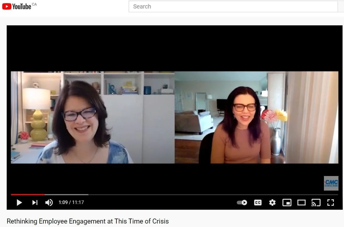Two women with dark hair and glasses smiling during a video call, one sitting in a room with a bookshelf and a lamp, and the other in a room with a vase of pink and yellow flowers, and a lamp.