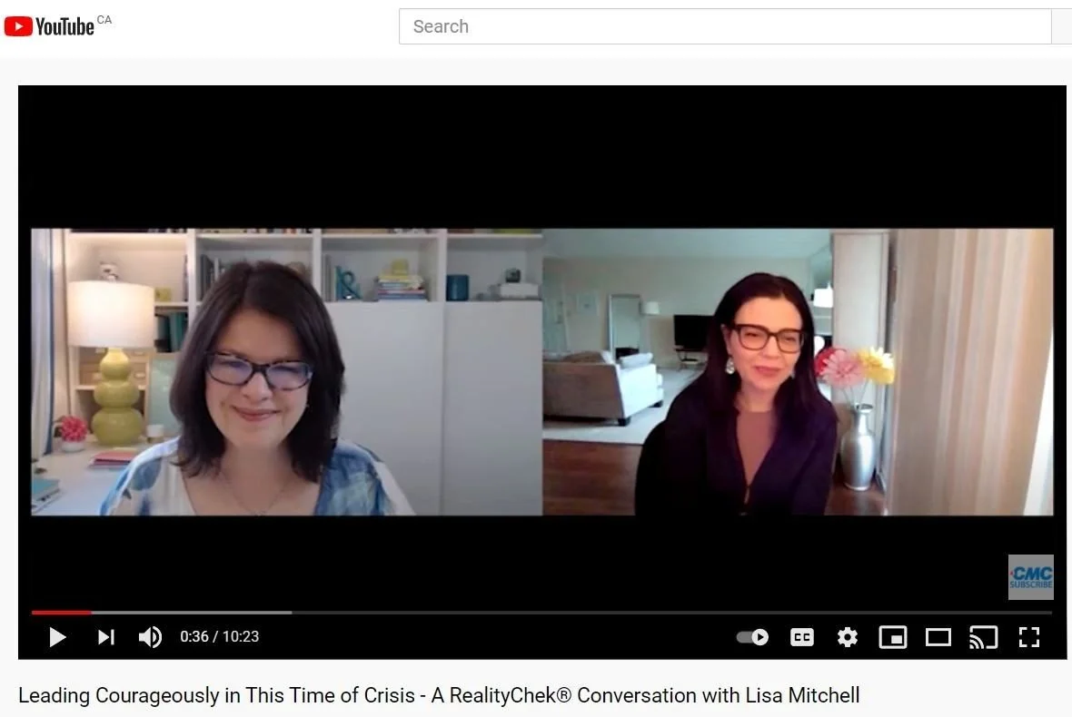 Two women participating in a virtual video call, each in their own home. The woman on the left has shoulder-length dark hair, glasses, and is smiling, with a well-lit background featuring shelves and a yellow lamp. The woman on the right has dark hair, glasses, and is also smiling, with a background showing a living room with a sofa, a small table with a lamp, and a vase of flowers.