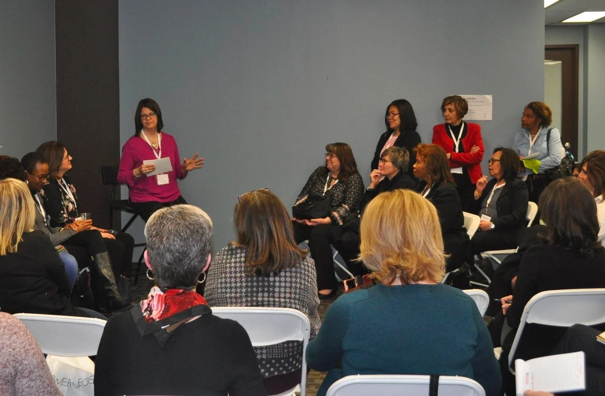 A woman in a pink top and black pants stands in front of a group of seated women, speaking and gesturing with her hand at a conference or seminar.