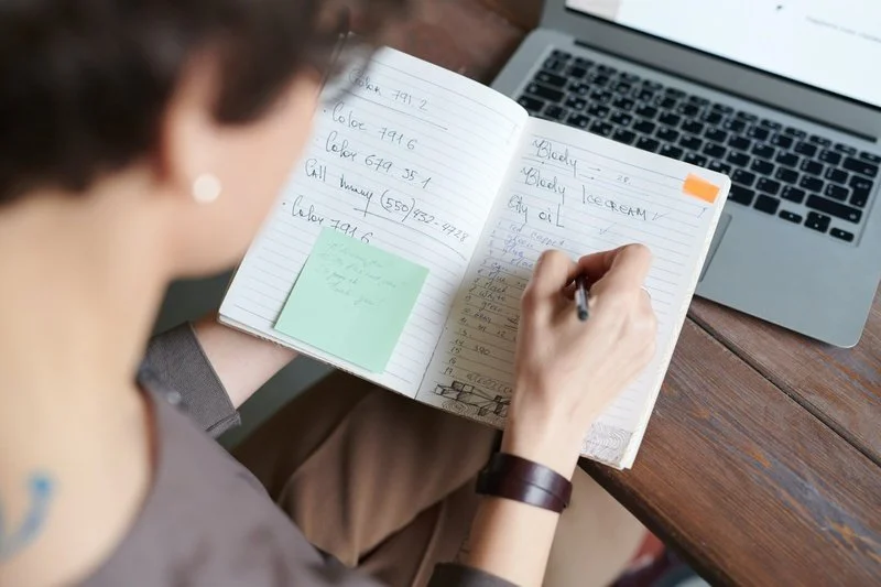 A person writing in a notebook at a wooden desk with a laptop nearby.