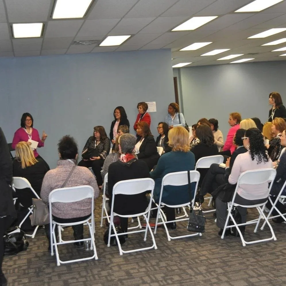 A group of women attending a conference or seminar, some seated and some standing, in a room with blue walls and fluorescent lighting.