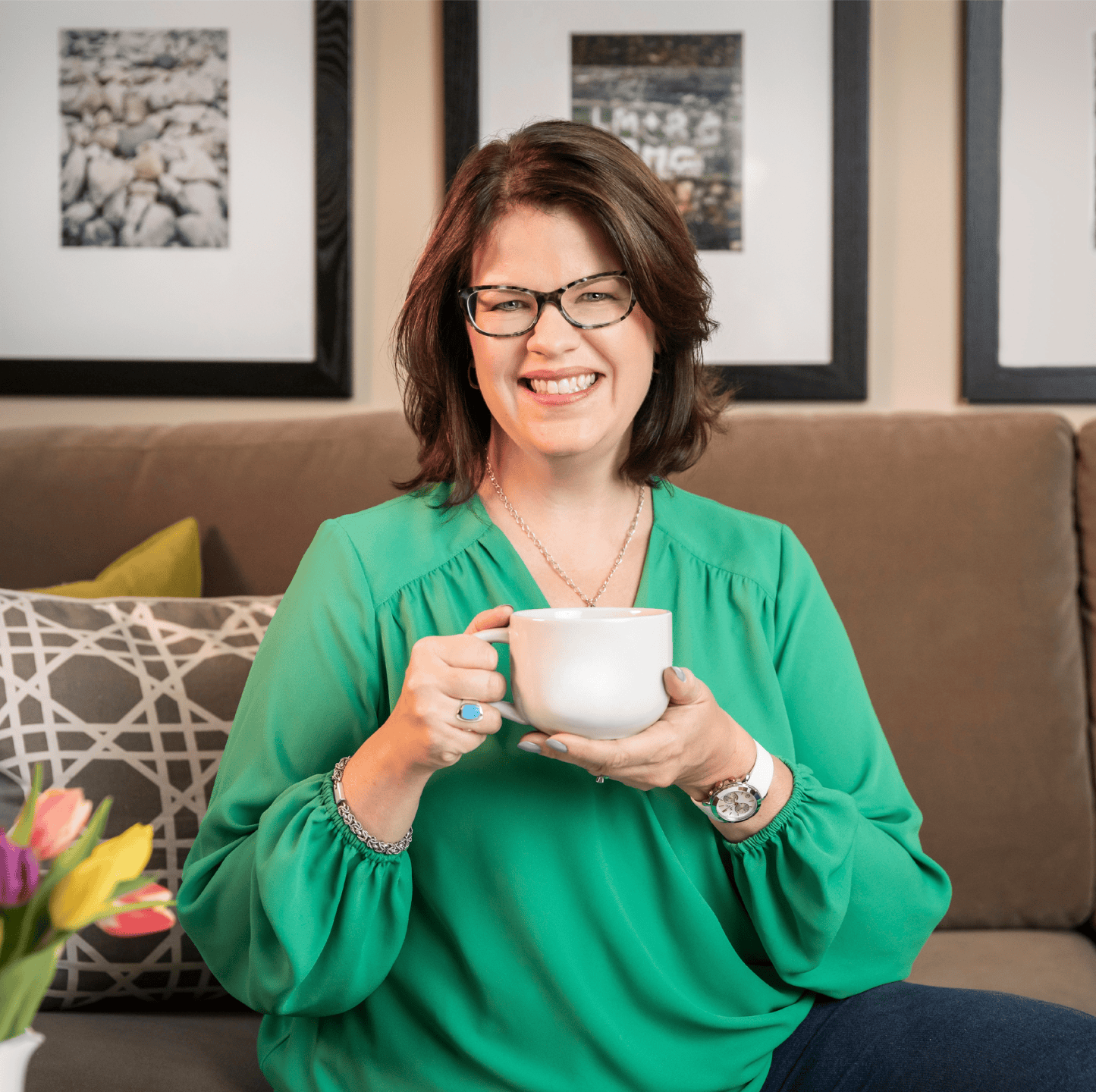 A woman with brown hair and glasses smiling while holding a white coffee mug. She is wearing a green blouse, sitting on a sofa with patterned and solid colored pillows, in a room with framed artwork on the wall.