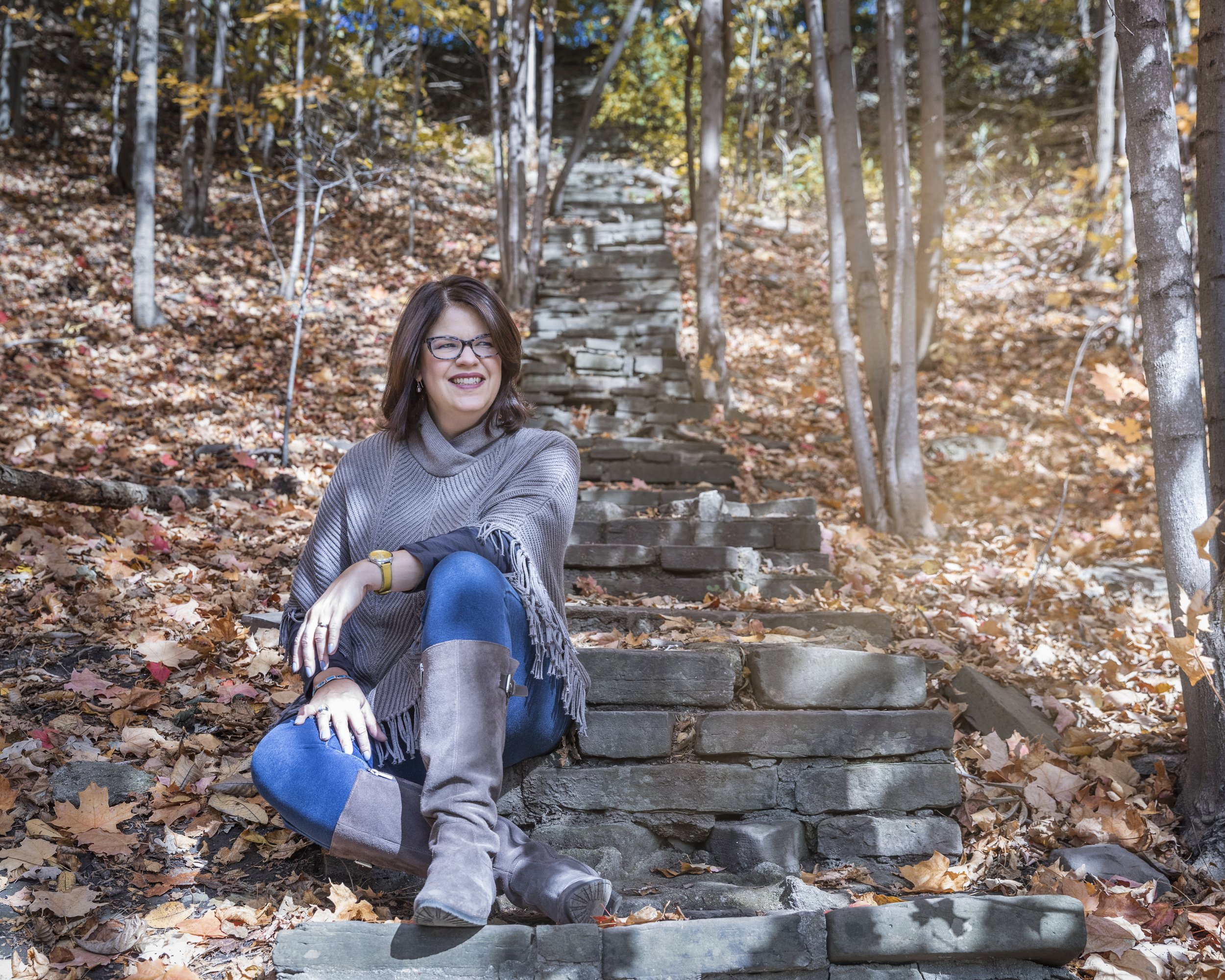 A woman with glasses sitting on stone steps in a wooded area with autumn leaves, smiling and wearing a gray sweater and boots.