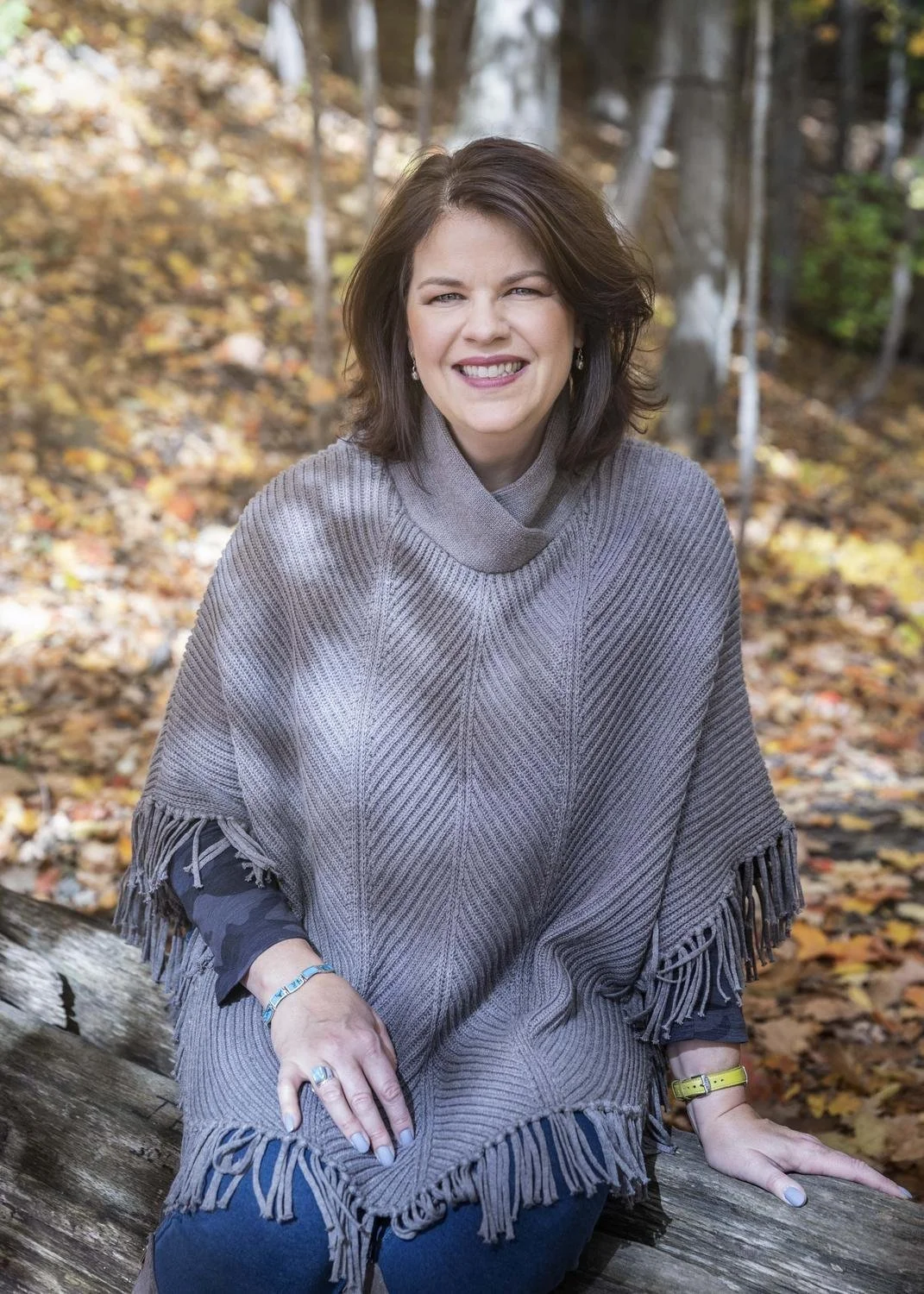 A woman sitting outdoors on a log in a wooded area with fallen autumn leaves, smiling at the camera.