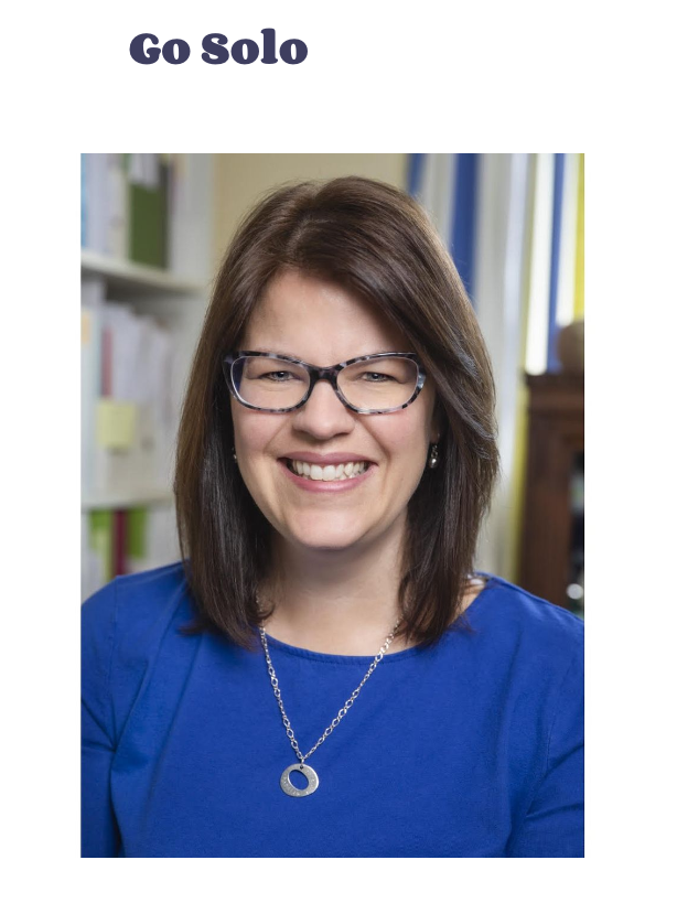 Smiling woman wearing glasses, a blue top, and a necklace, seated in an office or library with shelves of files behind her. Text at the top says "Go Solo".