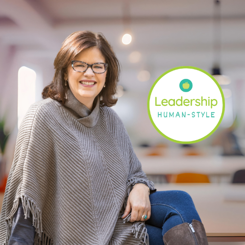 Woman with glasses smiling in an office, with a sign that reads 'Leadership Human-Style' and an apple icon.