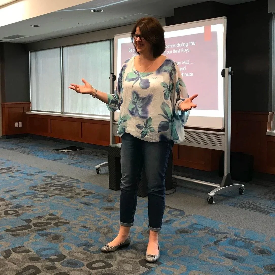 A woman with dark brown hair, glasses, and a floral blouse stands in front of a presentation screen during a talk or workshop in a conference room.