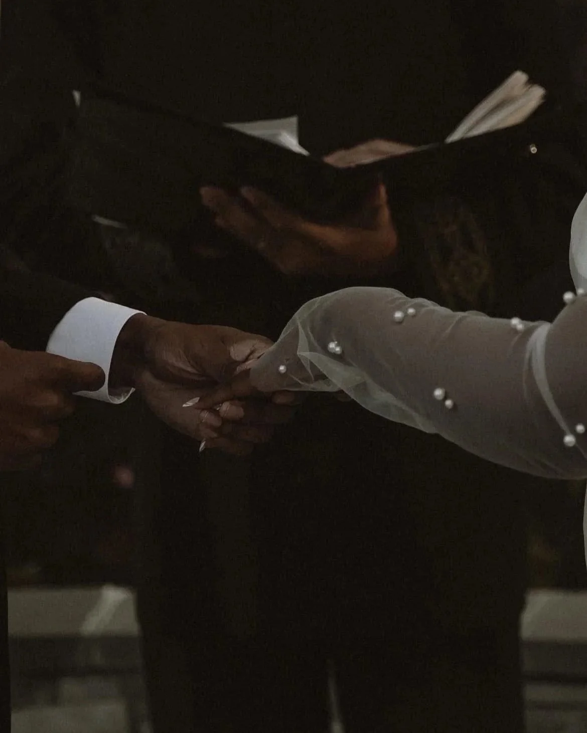 Close-up of a couple holding hands during a wedding ceremony, with one person wearing a white sleeve with pearl embellishments and the other in black attire, in a dark setting.