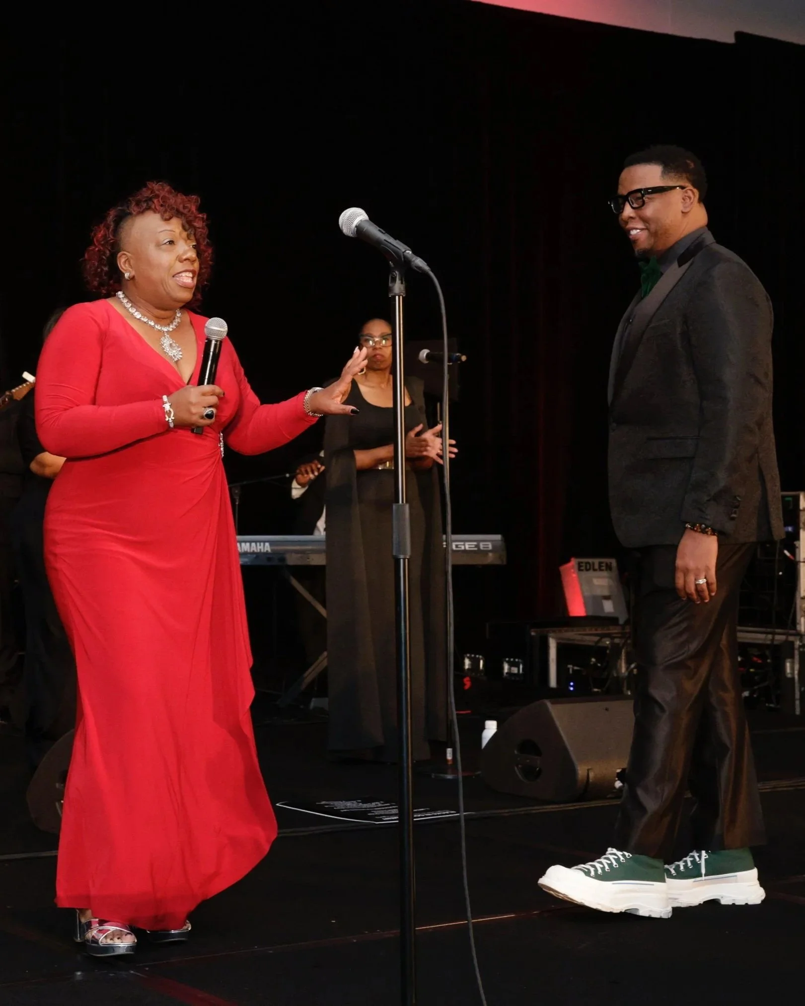 Woman in red dress and jewelry speaking into microphone to man in black suit with green shoes on a stage with musical equipment and a woman in black in the background.