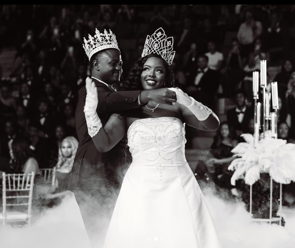 A black-and-white photo of a bride and groom dancing at their wedding reception. The groom, wearing a crown, is embracing the bride, who is wearing a crown and a strapless wedding dress. They are smiling, and there are guests in the background, along with a decorative table with candles.