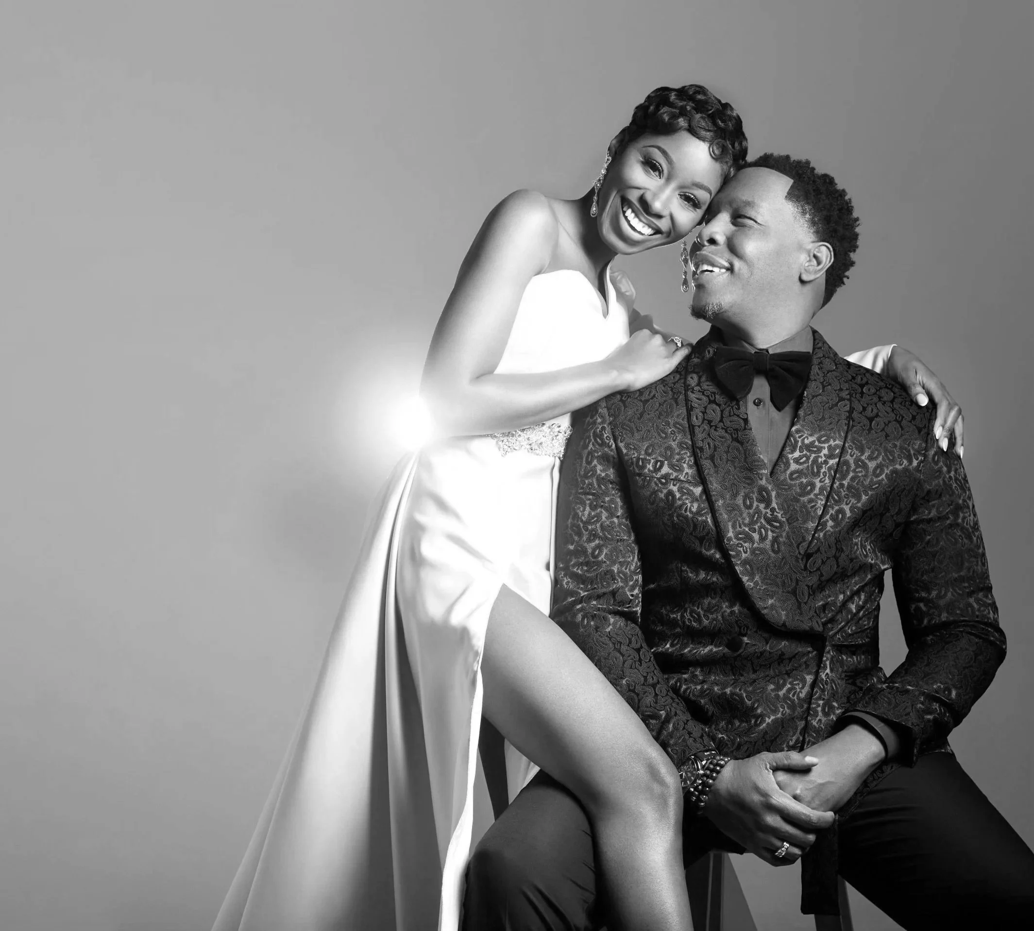 A black-and-white portrait of a smiling woman in a wedding dress and a man in a tuxedo with a bow tie posing together, with the woman standing and the man seated, both exuding joy and affection.