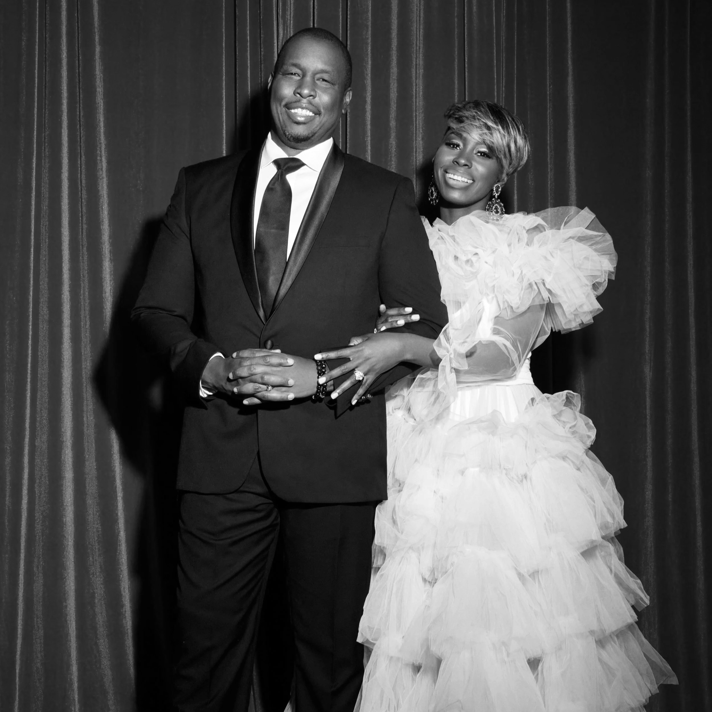 Black and white photo of a smiling man in a tuxedo and a smiling woman in a formal gown, standing together against a curtain backdrop.