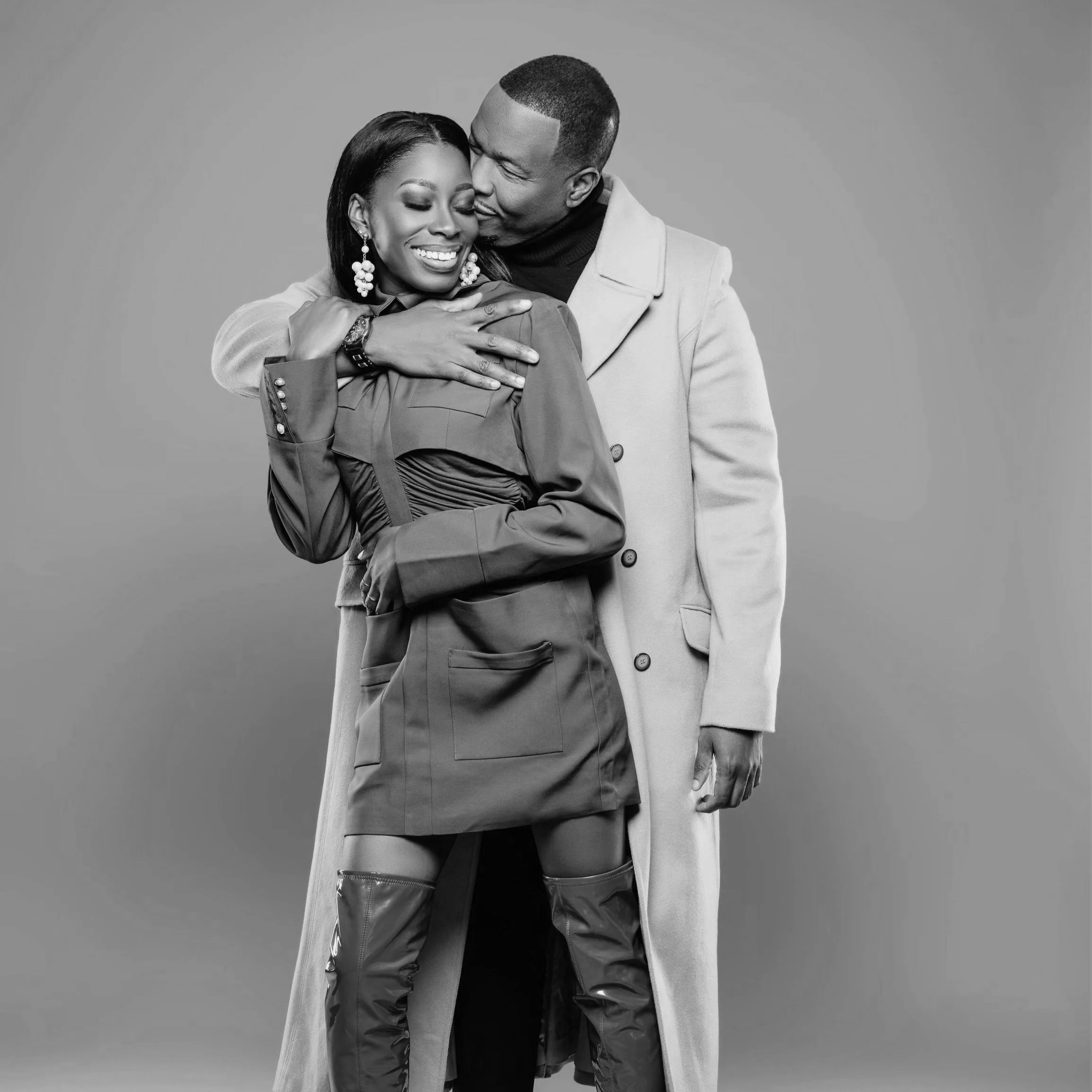 A black and white photo of a smiling woman with long earrings, wearing a coat, being embraced from behind by a man in a coat, both showing affection.