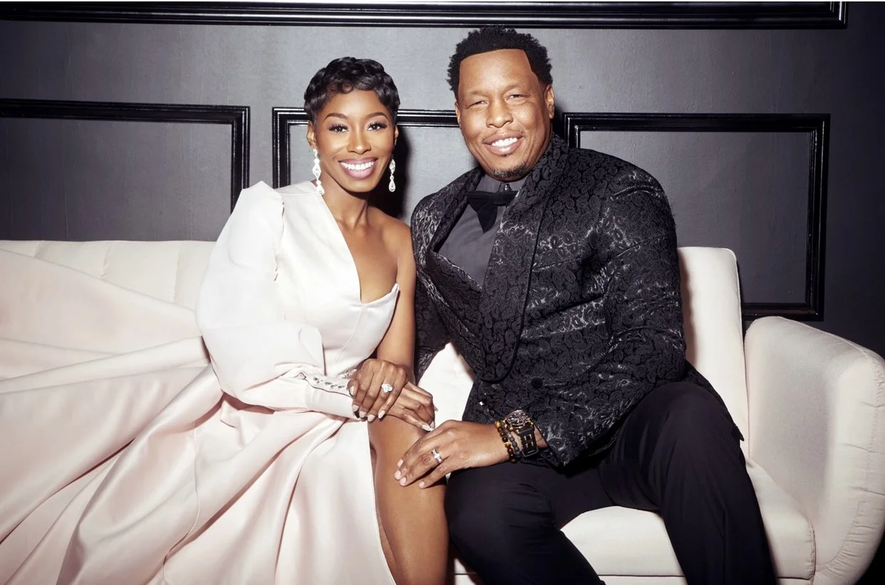 A smiling woman in a white dress and a smiling man in a black patterned blazer, sitting together on a white sofa in a dimly lit room with dark walls.