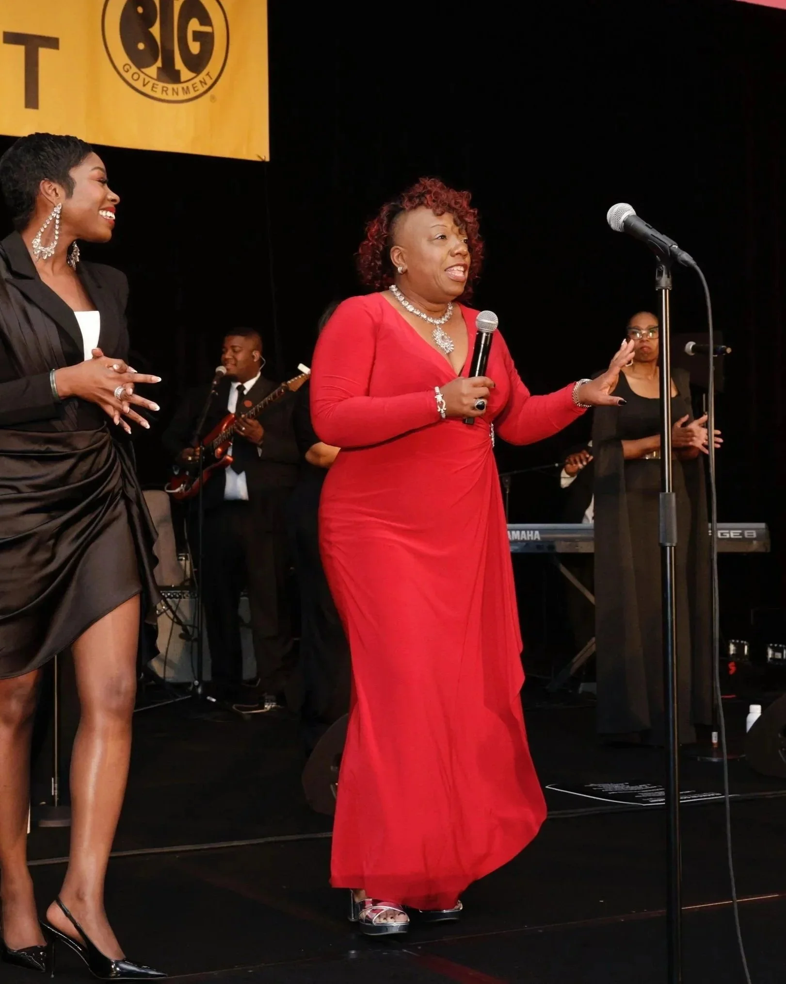 A woman in a red dress speaking into a microphone on stage, with other formally dressed musicians and singers in the background.