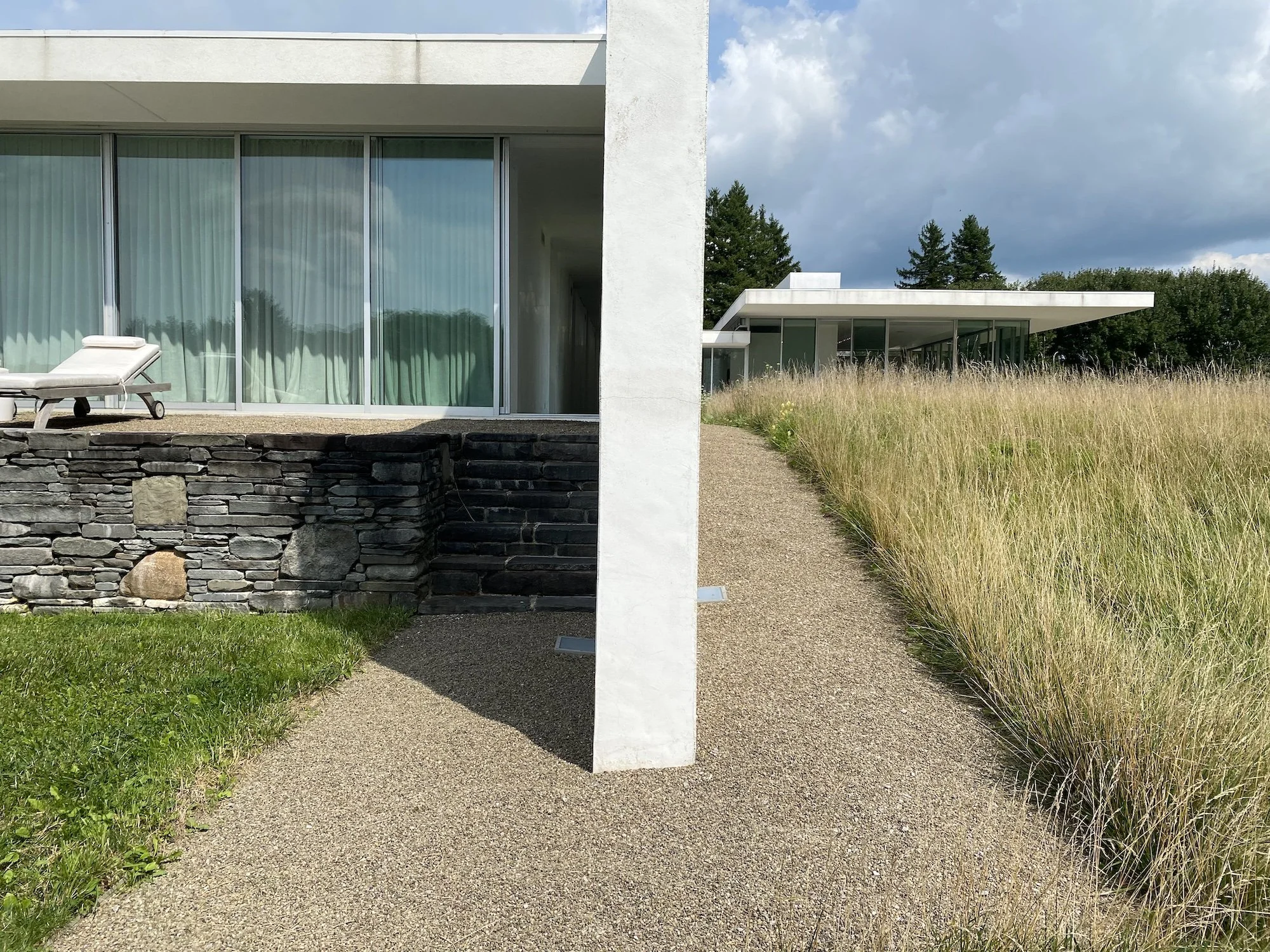 Gravel path beside Fairlawn’s glass facade, stone terrace, and tall meadow grasses.