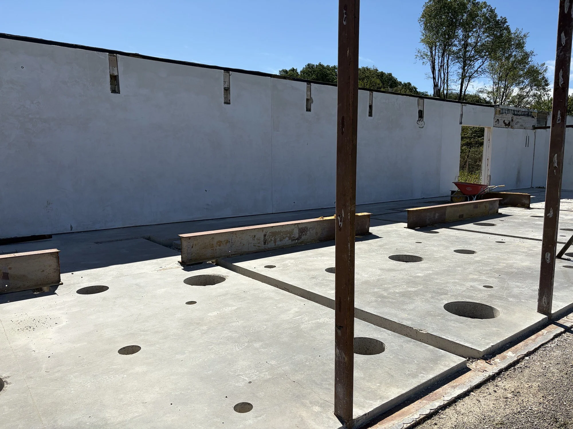 Exposed concrete floor with circular openings, steel columns, and a retained white wall.