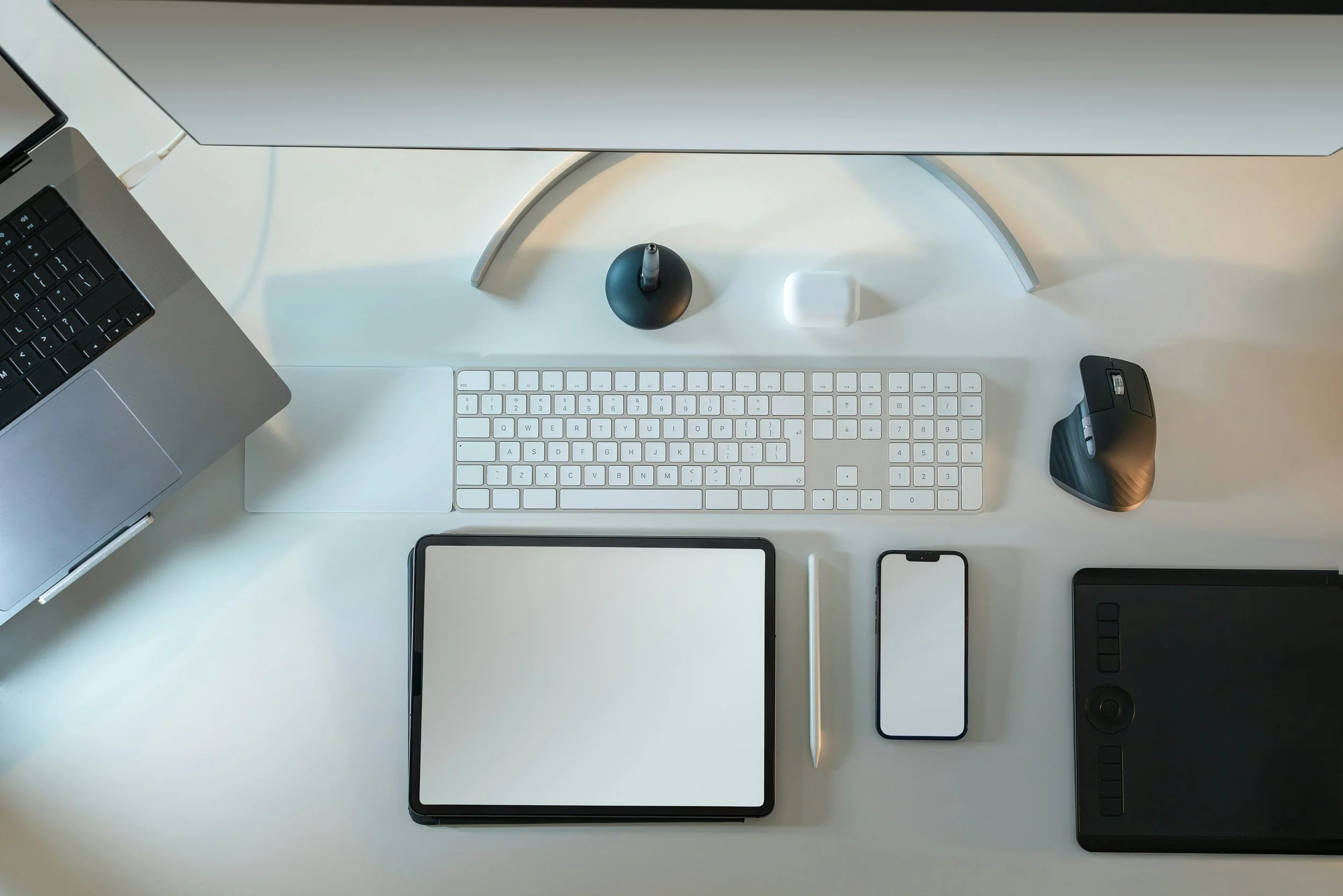 Overhead view of a modern workspace with a monitor, keyboard, stylus, tablet, smartphones, and a wireless mouse on a white desk.