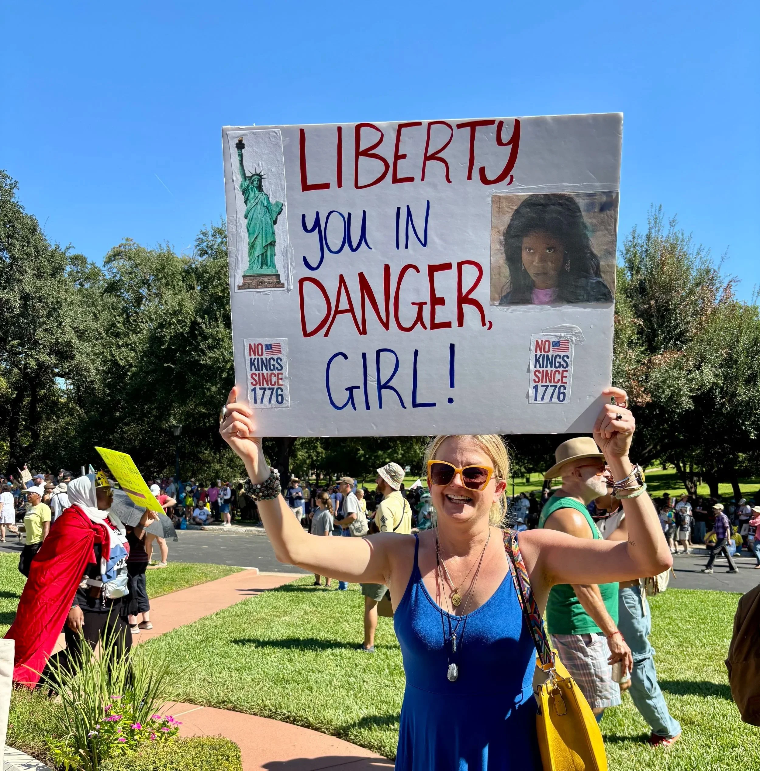 A woman holding a protest sign with an image of the Statue of Liberty, a photograph of a girl, and slogans like "Liberty, You in Danger, Girl!" during a public demonstration, with a crowd of people in the background.