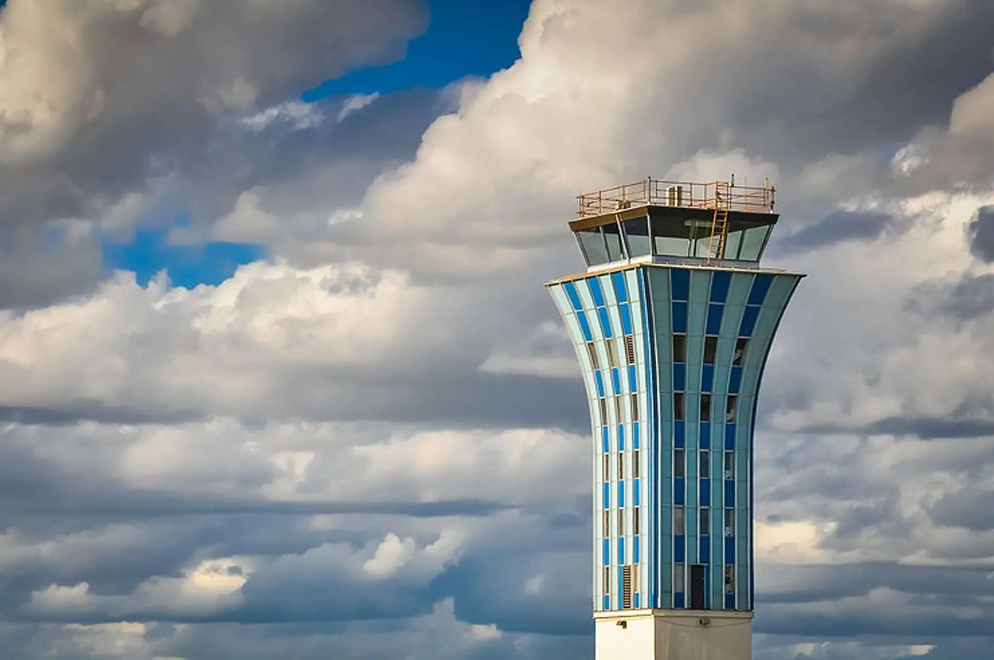 The image shows an airplane control tower with a cloudy sky in the background. The tower has a modern design with blue and white panels and a glass-enclosed top observation area.