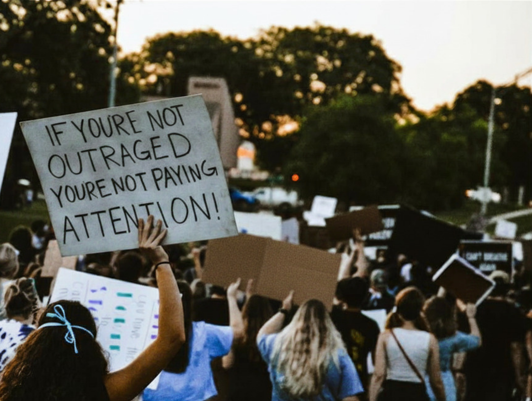 A crowd of people protesting outdoors during sunset, with one person holding a sign that reads, 'IF YOU'RE NOT OUTRAGED YOU'RE NOT PAYING ATTENTION!'