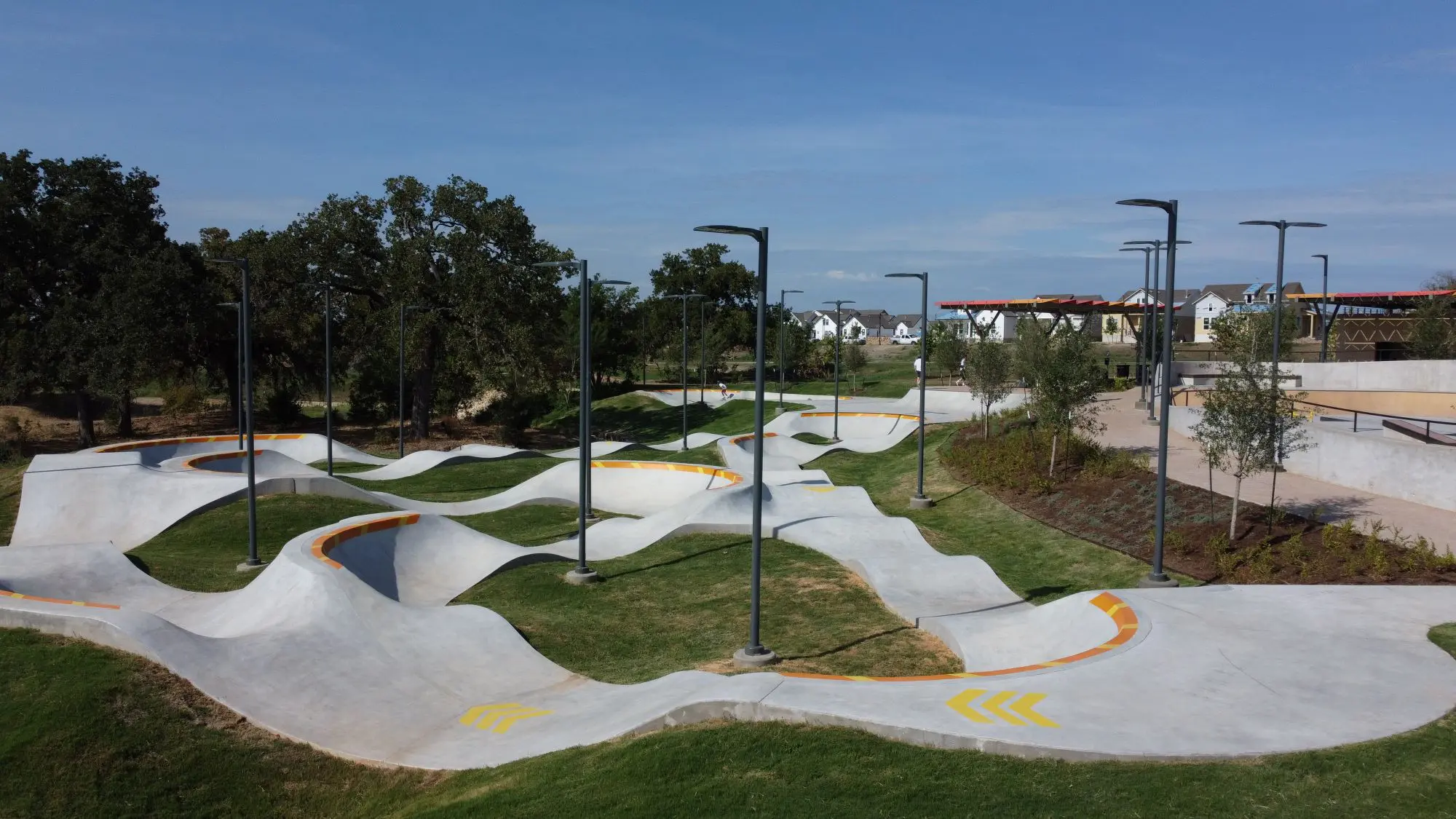 A skatepark with white concrete ramps and bowls, surrounded by small trees and street lamps under a blue sky, with residential buildings in the background.