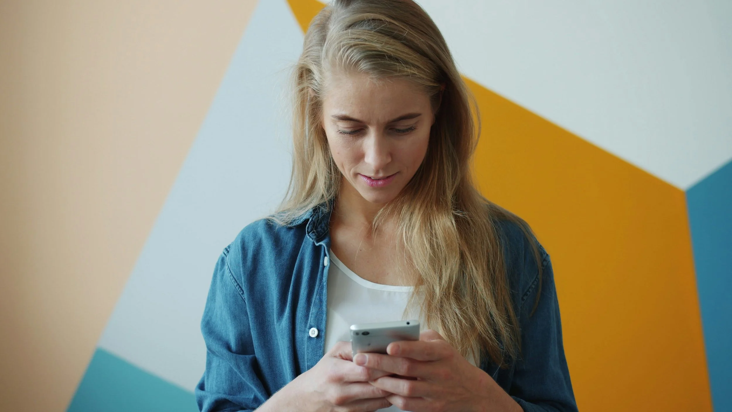 A young woman with long blonde hair looking down at her phone, wearing a denim jacket over a white shirt, standing against a colorful geometric wall.