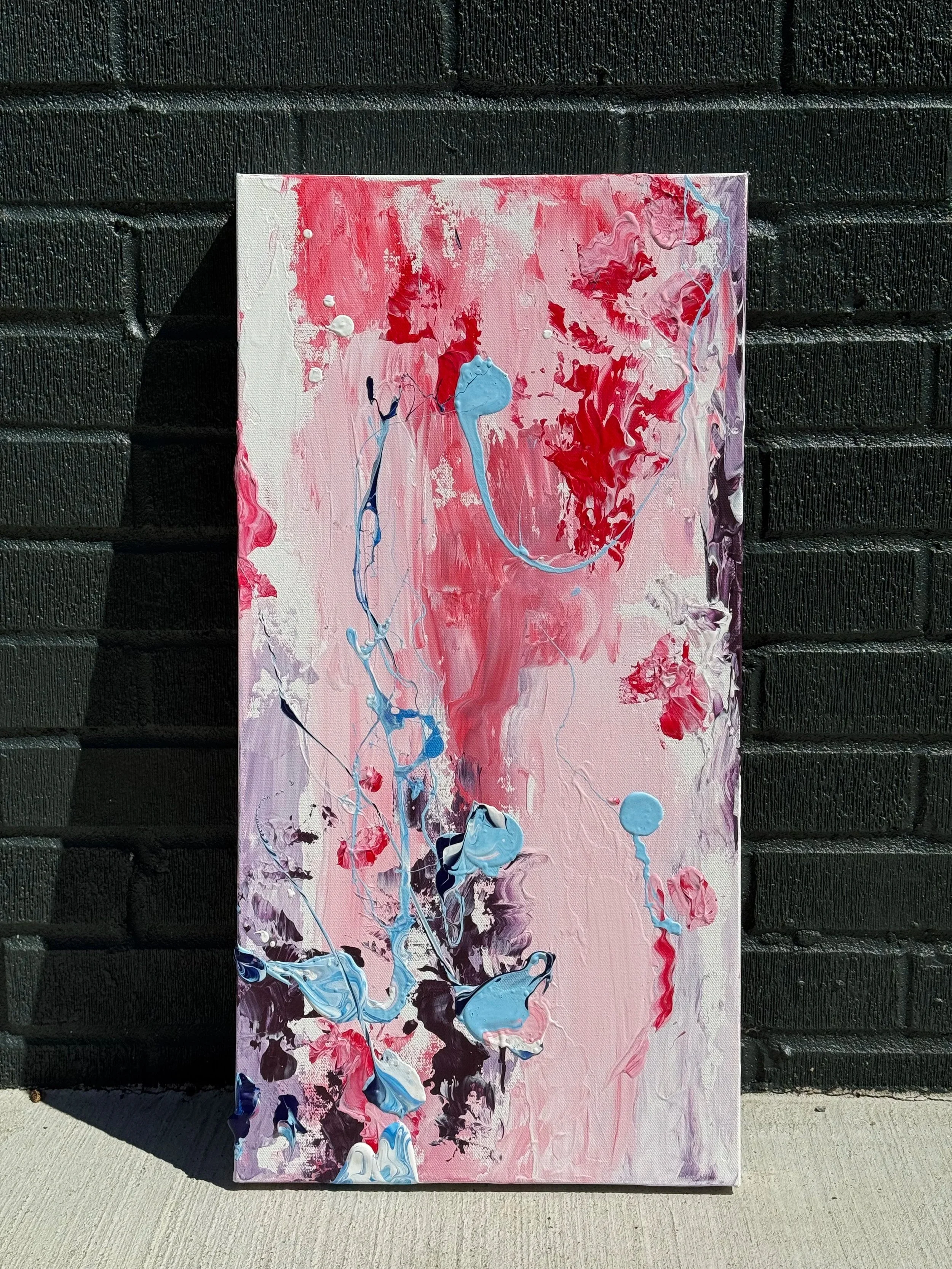 Abstract painting with pink, red, blue, purple, and black paint splatters and streaks, leaning against a black brick wall outdoors.
