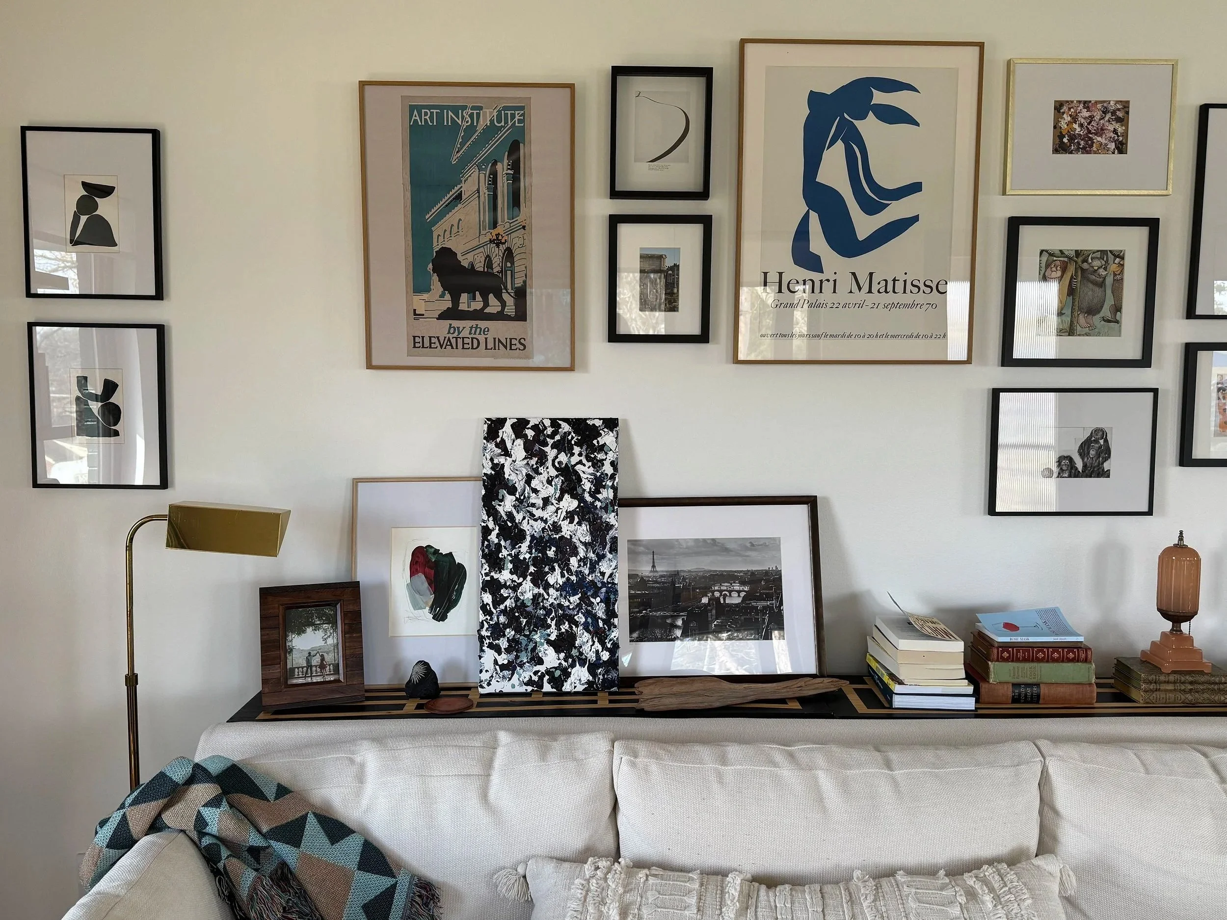 Photo of a wall with multiple framed artworks, including posters and prints, above a white sofa. The decor includes a lamp, books, and personal items on a shelf along the sofa. A patterned blanket is draped over the arm of the sofa.