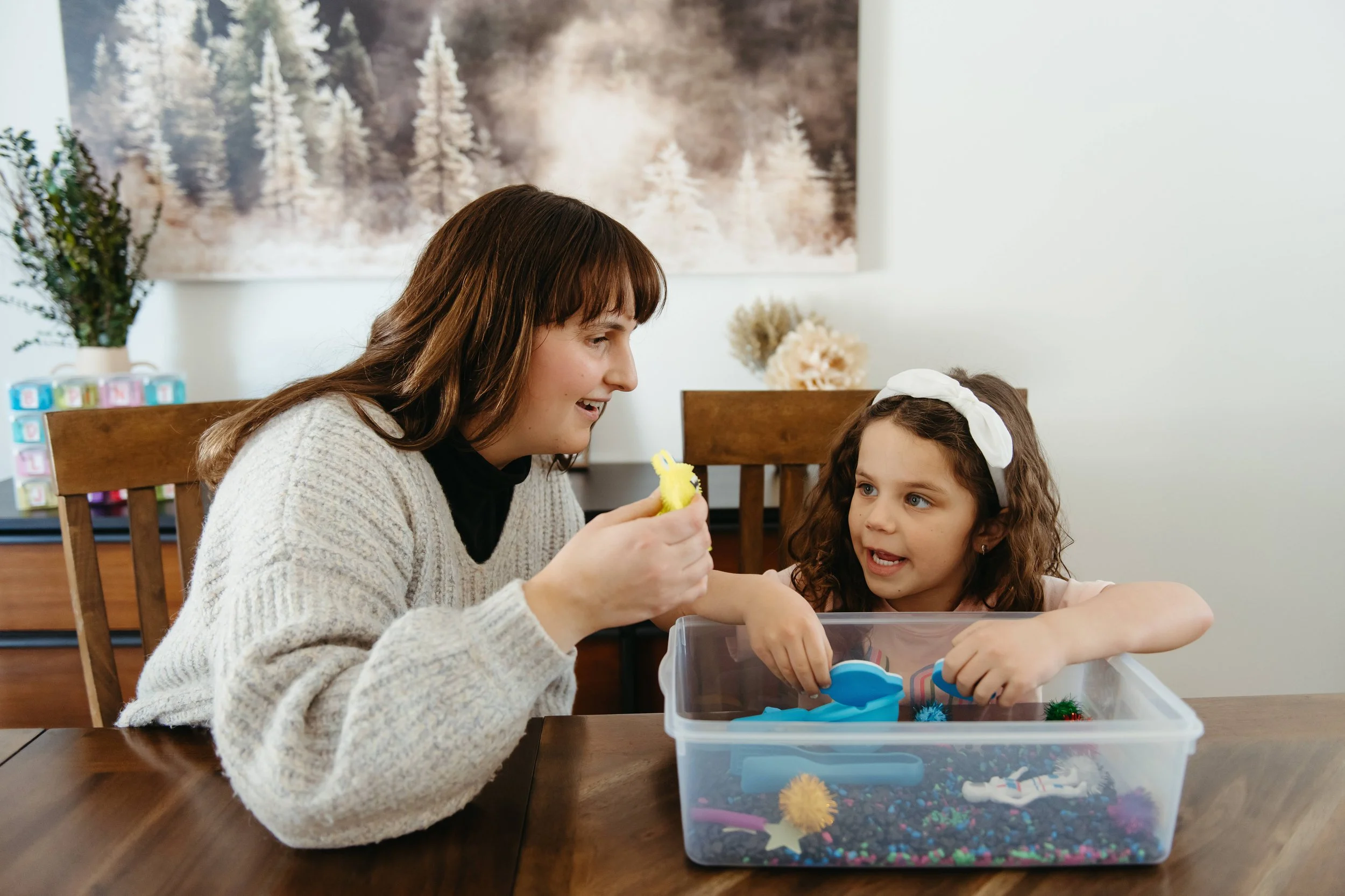 A woman and a girl playing with sensory toys at a wooden table in a room decorated with a winter-themed picture and plants.