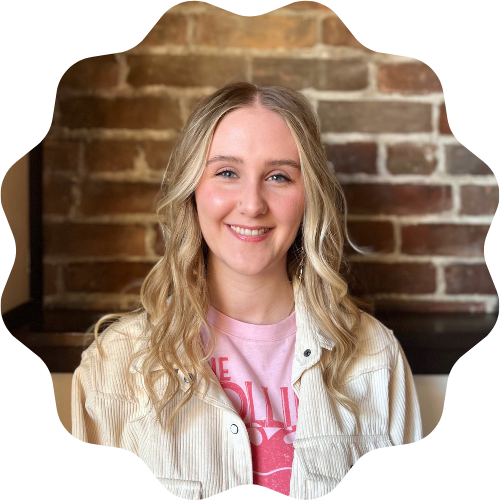 A woman (Kaelyn McCallum) with blonde hair smiling, wearing a light pink T-shirt and a beige jacket, standing in front of a brick wall.