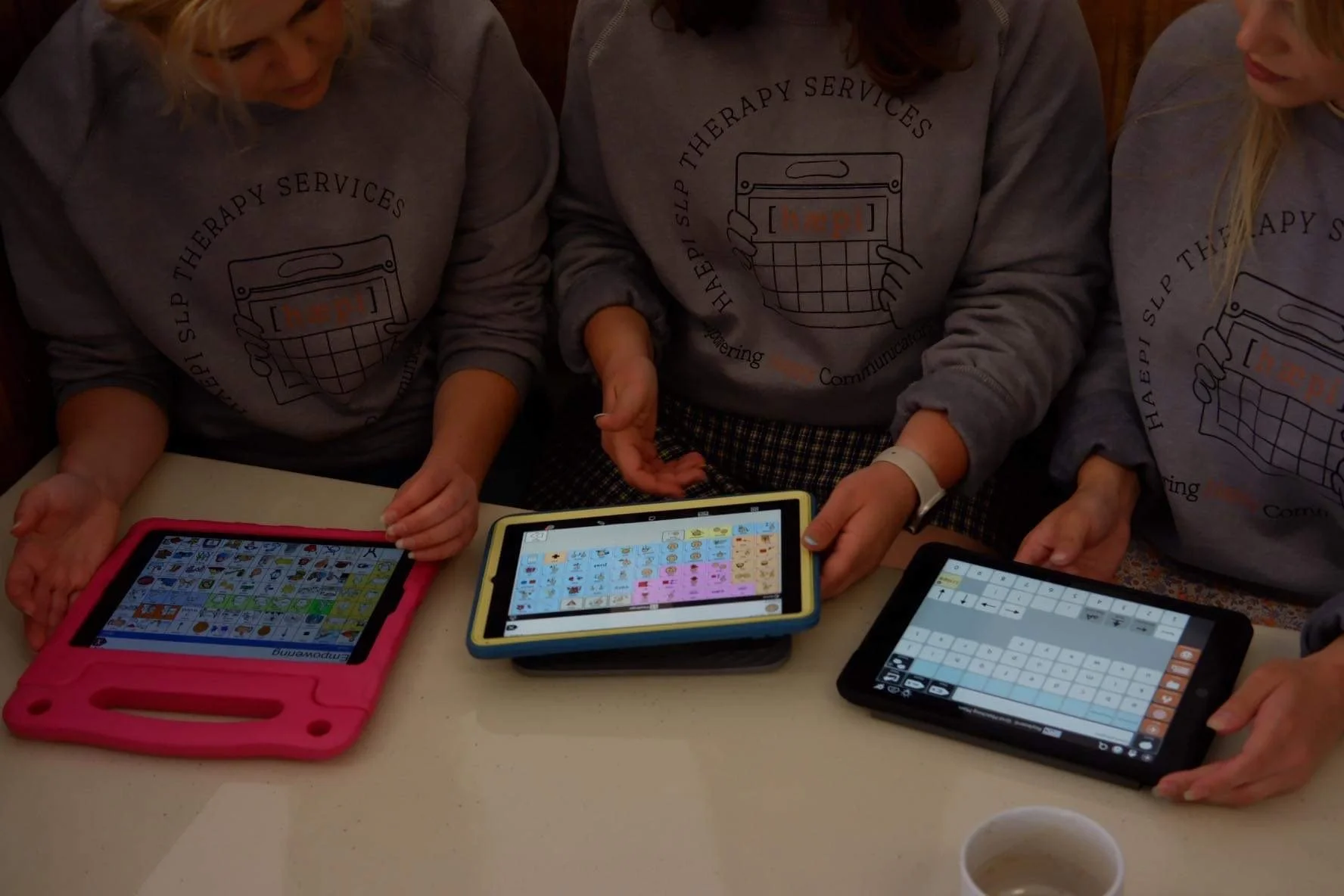 Three people wearing gray sweatshirts with the HAEPI SLP Therapy Services logo, sitting at a table with three tablets displaying alternative and augmentative communication apps.
