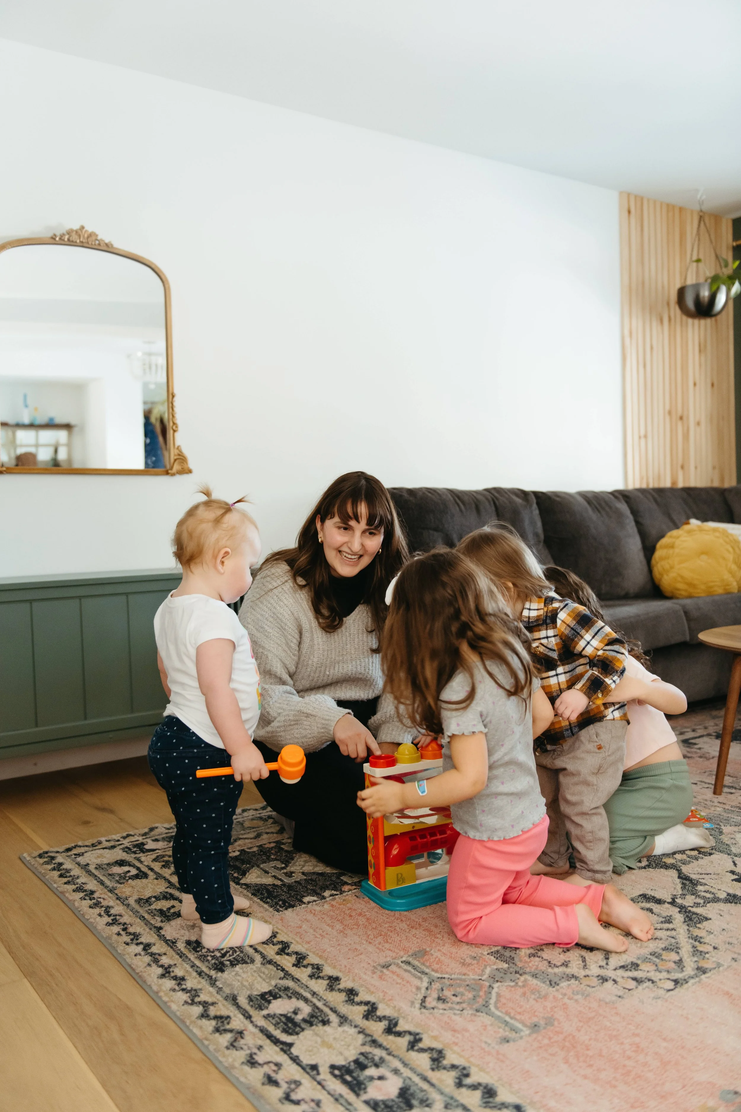 An SLP and four children playing together in a living room with hardwood floors, a patterned rug, a gray couch, and a mirror on the wall.
