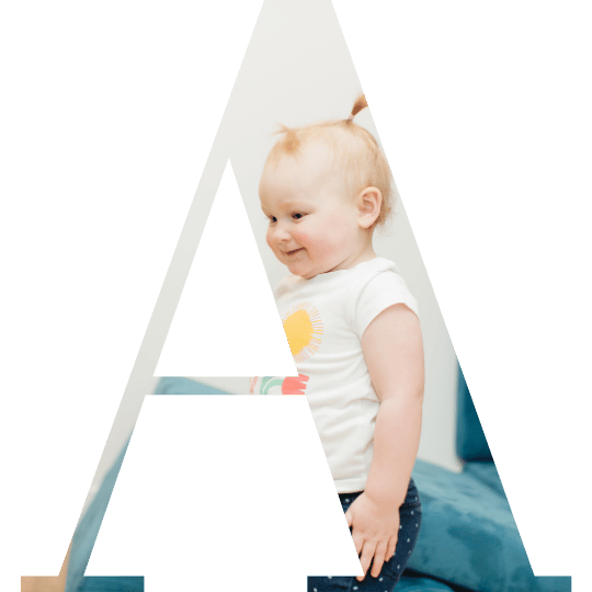 A toddler girl with blonde hair and a white t-shirt, sitting on a bed or couch.