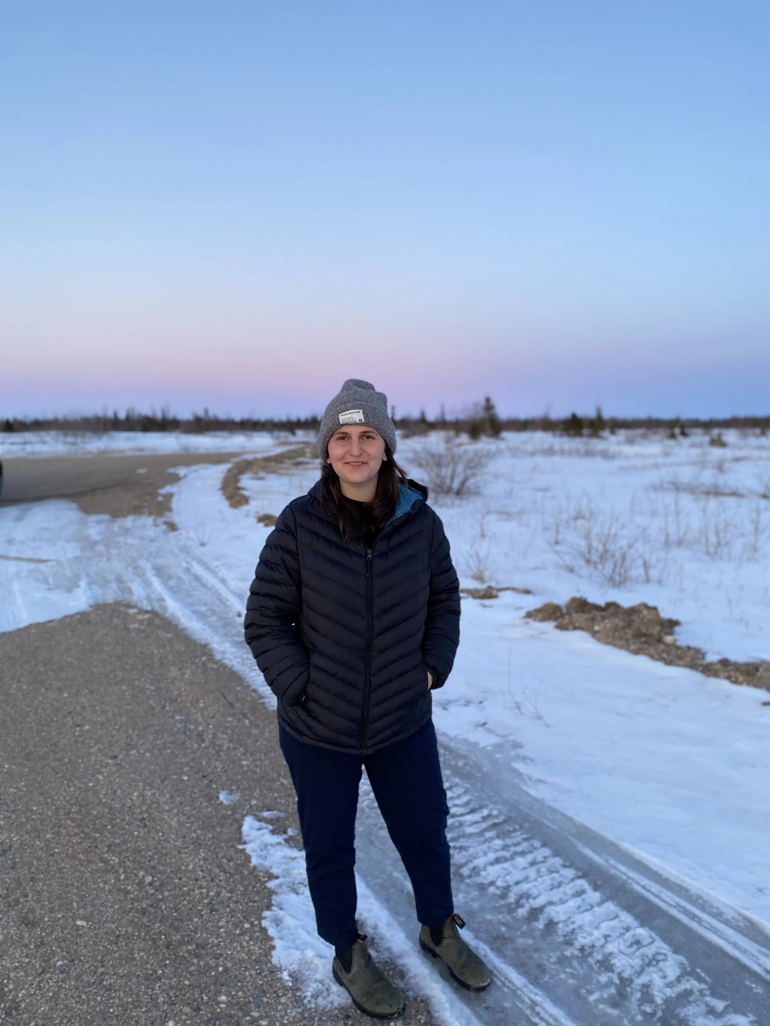A woman (Halle Demchuk) standing on a snowy road at dusk, wearing a gray beanie, black puffer jacket, and dark pants, with a landscape of snow and sparse trees in the background.