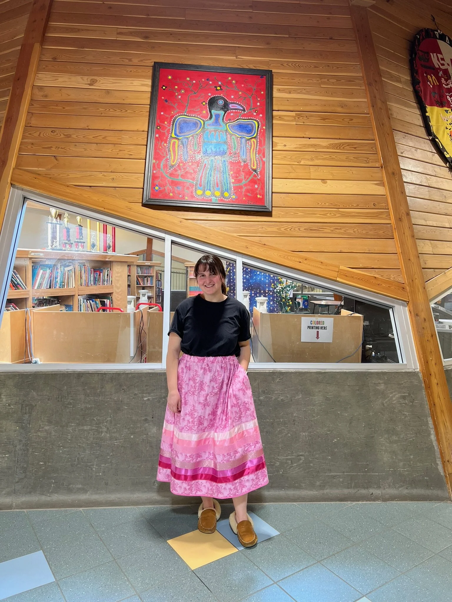 A woman (Halle Demchuk) wearing a black t-shirt, a pink ribbon skirt, and brown shoes standing indoors in front of a window. Behind her is a wooden wall with a colourful painting above the window. The interior includes bookshelves and trophies.