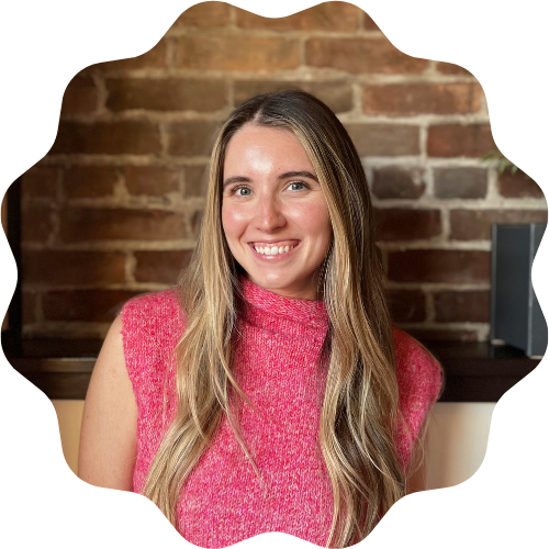 A woman (Megan Kane) with long blonde hair wearing a pink sleeveless sweater, smiling in front of a brick wall.