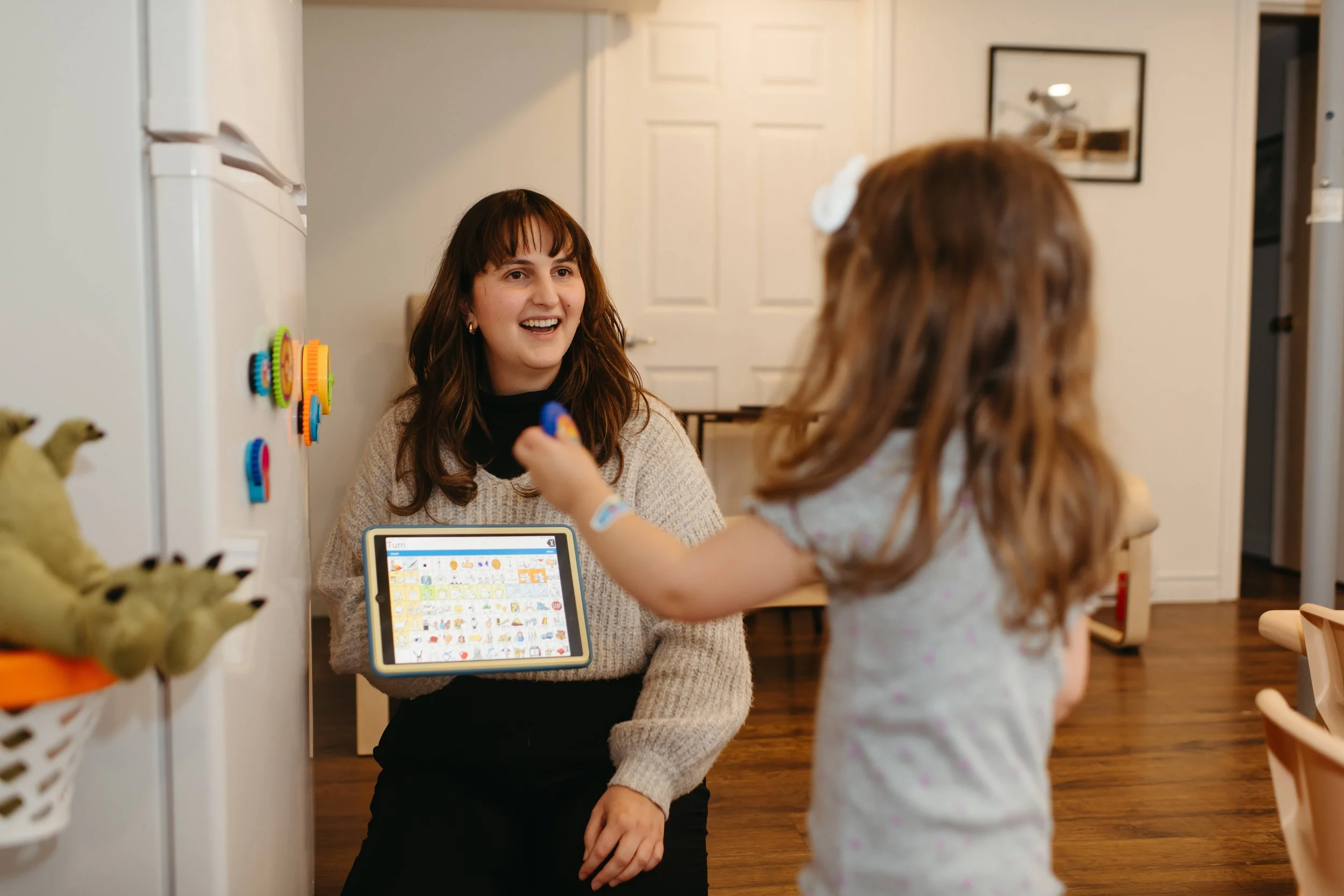 A young woman sitting on the floor, smiling and holding a tablet, while a little girl with a bow in her hair stands in front of her holding a toy. The scene takes place in a kitchen with wooden floors and kid-friendly decor.