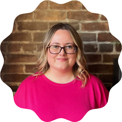 A woman (Megan Hutchinson) with blonde hair and glasses wearing a bright pink shirt, standing in front of a brick wall.