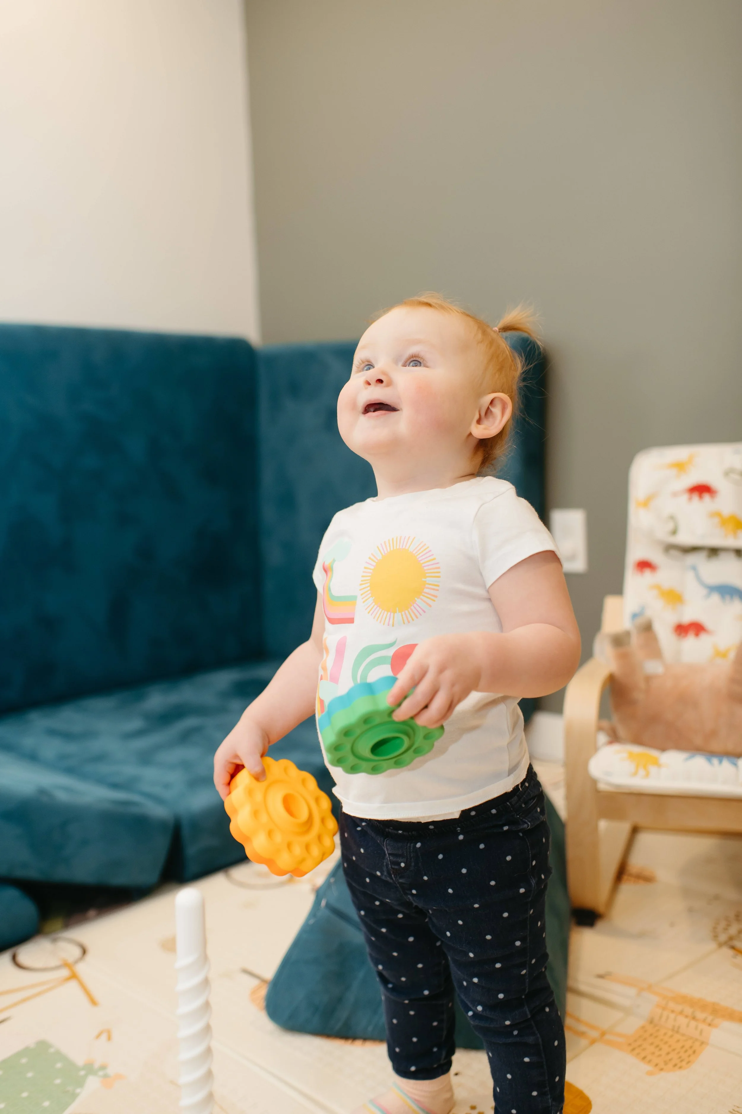 A young child with red hair and blue eyes stands indoors, holding two colorful textured toys, smiling and looking up.